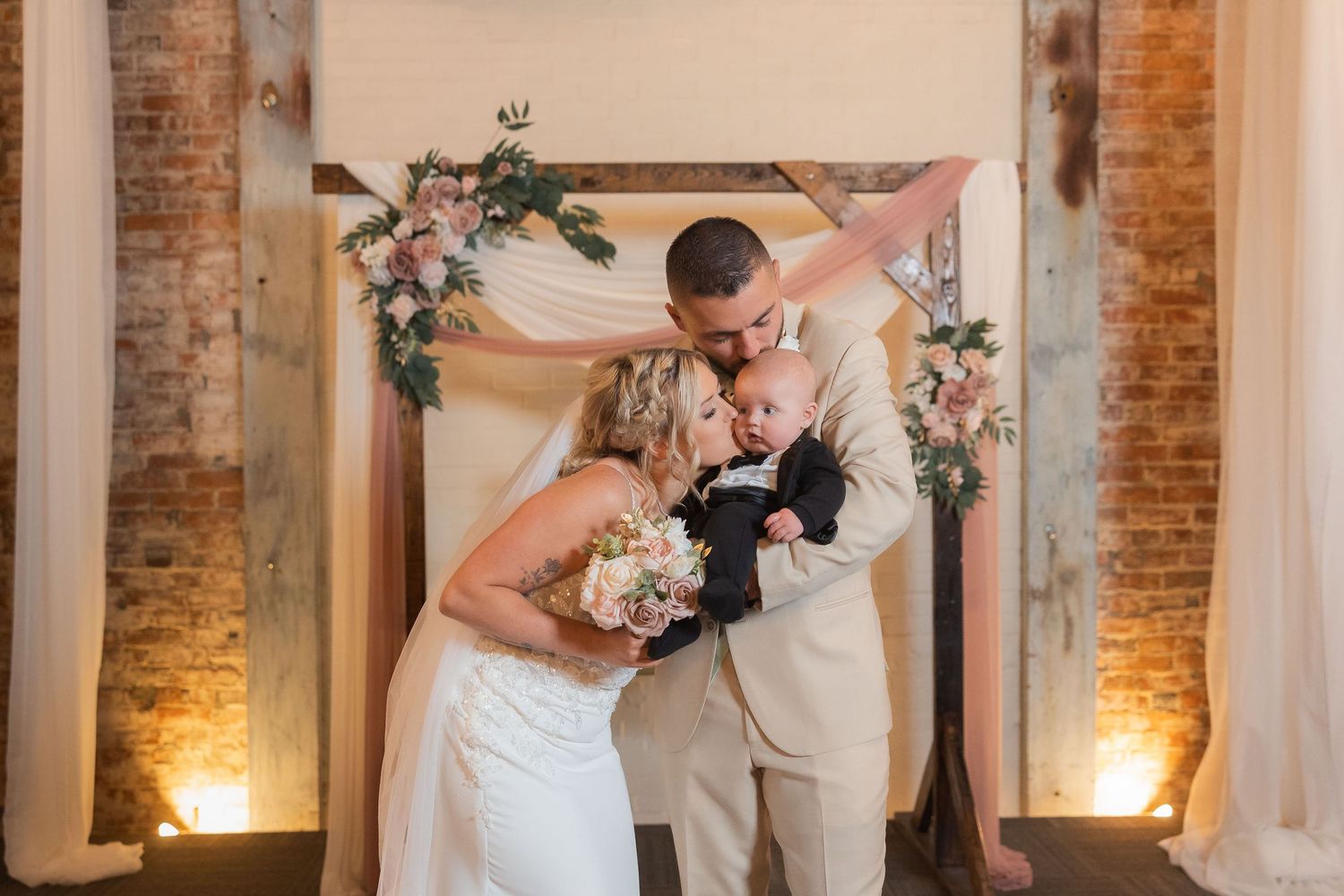 Wedding couple poses against exposed brick walls decorated with greenery and soft lighting.