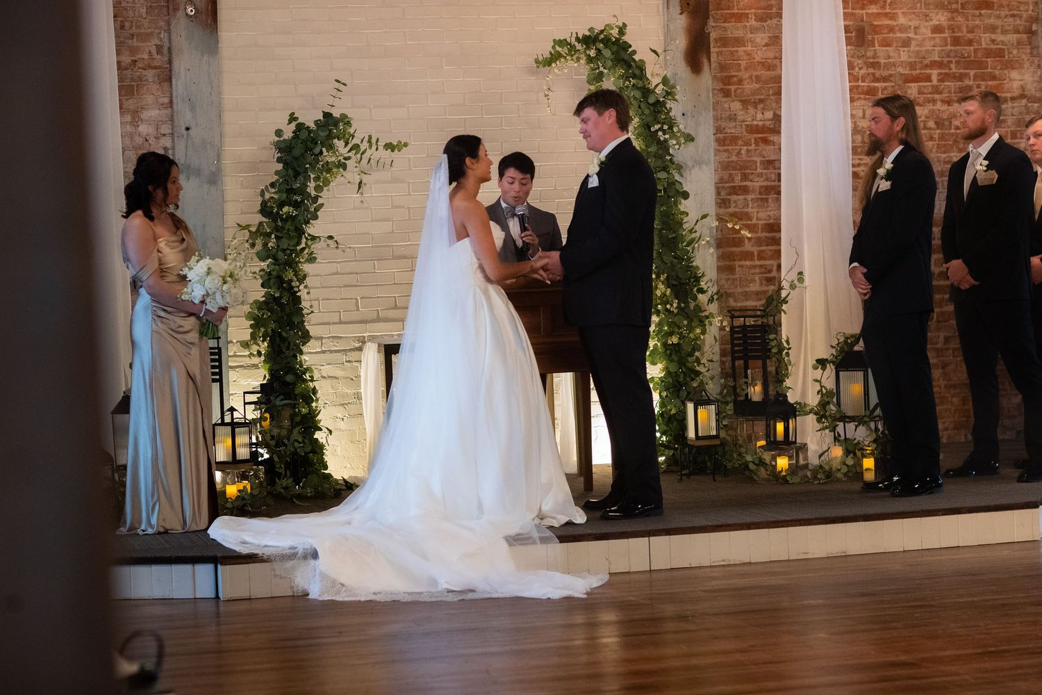 A wedding ceremony in progress at a rustic brick venue with greenery arch decorations.