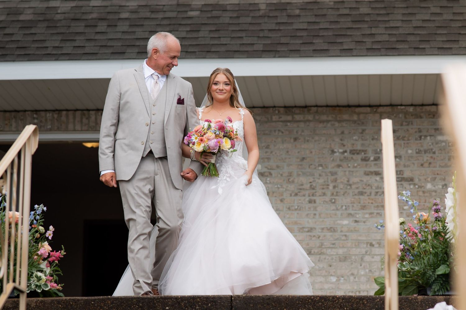Bride in white gown holding colorful bouquet walks with older man in gray suit down church steps.