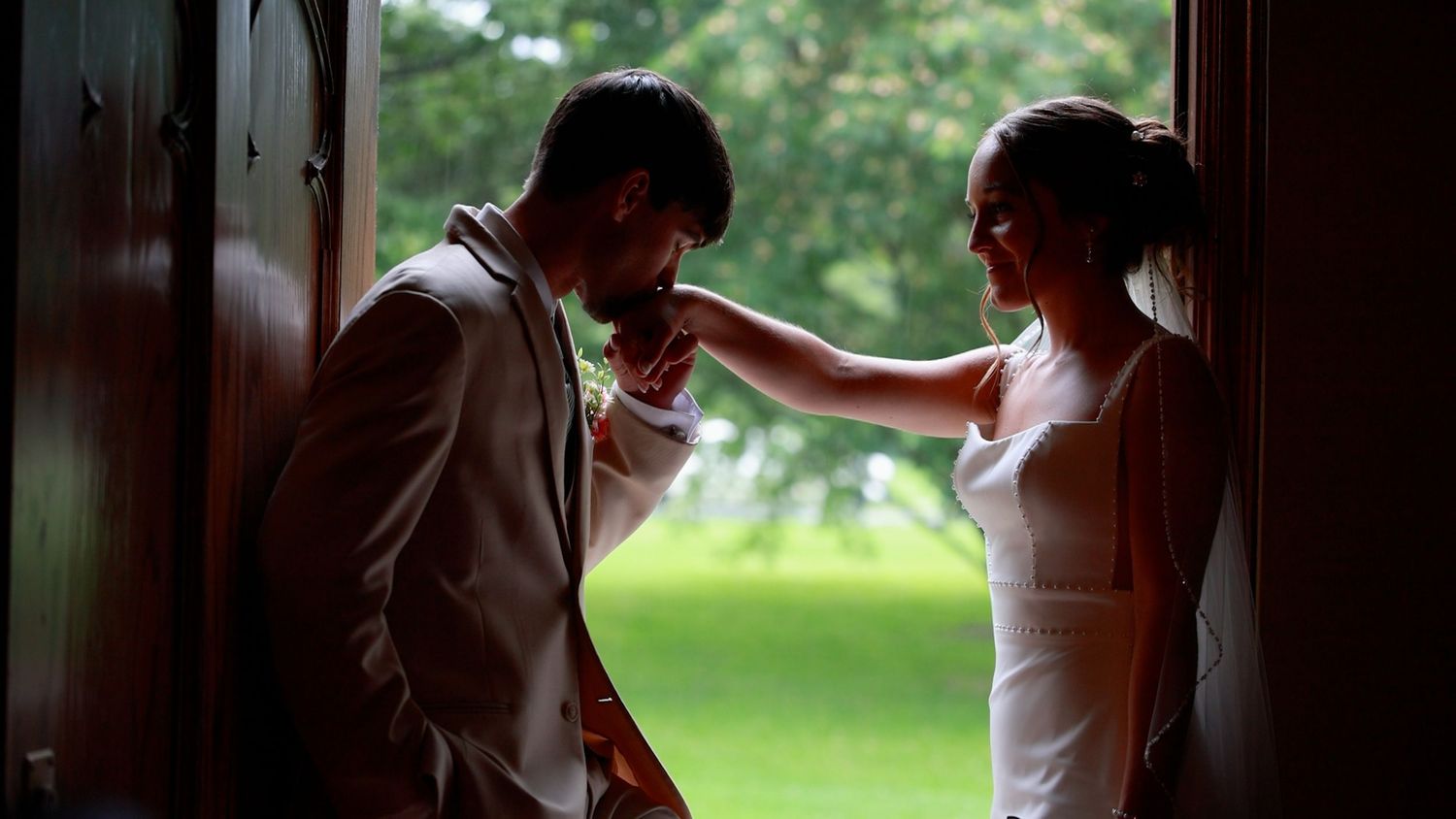 Bride adjusting groom's tie in a doorway with lush green garden background on wedding day.