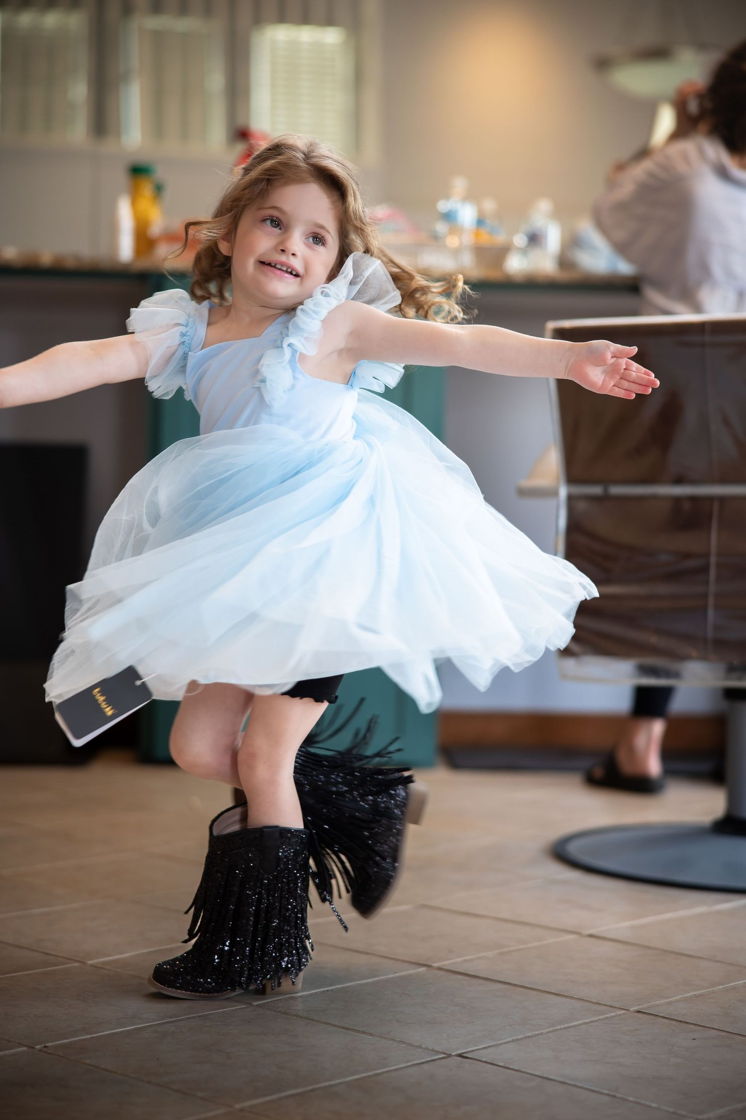 A young girl in a light blue princess dress and black fringe boots twirls joyfully indoors.
