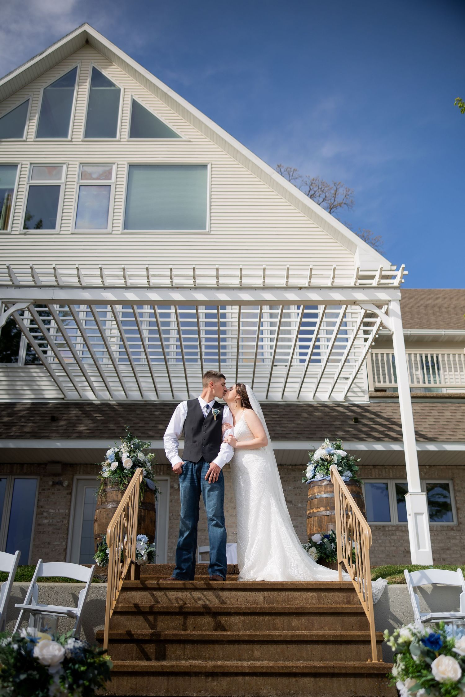 Newlyweds share a kiss on decorated staircase in front of elegant white venue with blue sky backdrop.