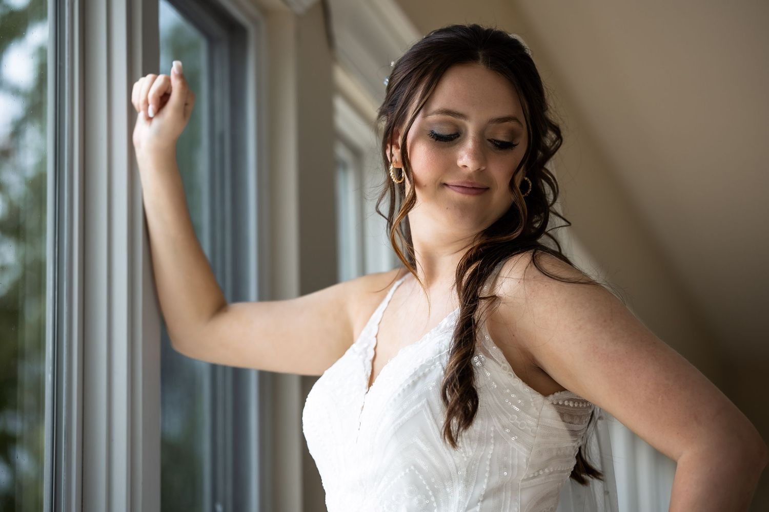Bride in white lace wedding dress standing by window, smiling softly with elegant updo hairstyle.