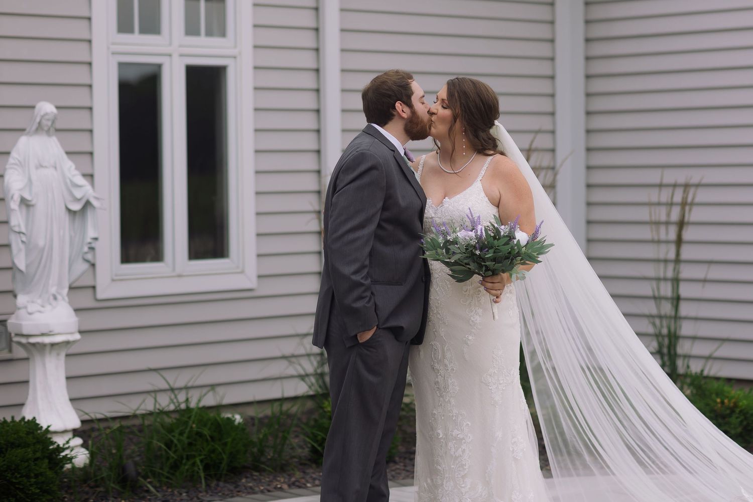 Bride and groom share a kiss outdoors near a white Virgin Mary statue, bride holding purple floral bouquet.