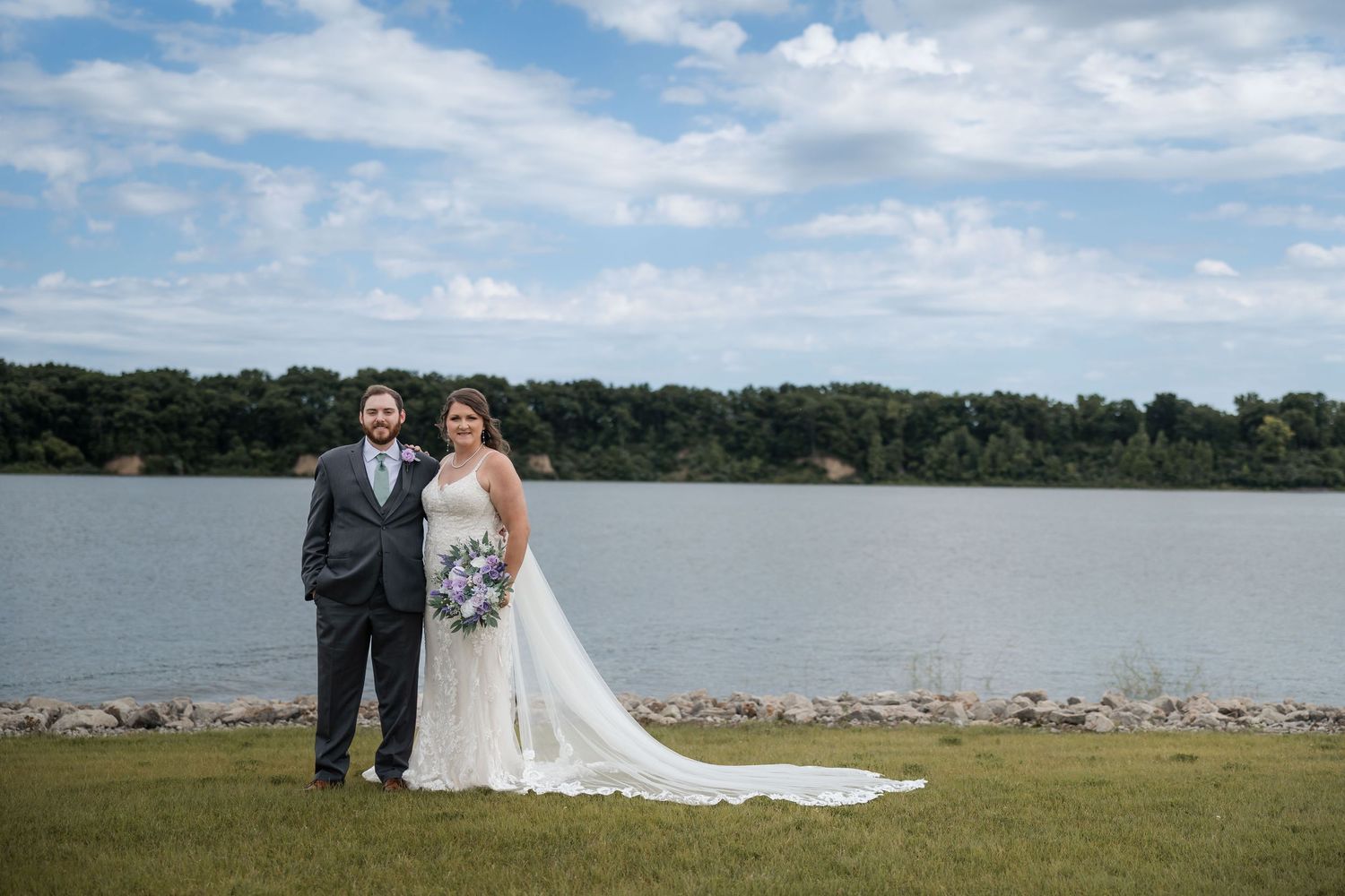 Bride and groom posing by a lakeside on their wedding day, bride holding purple bouquet in white gown.