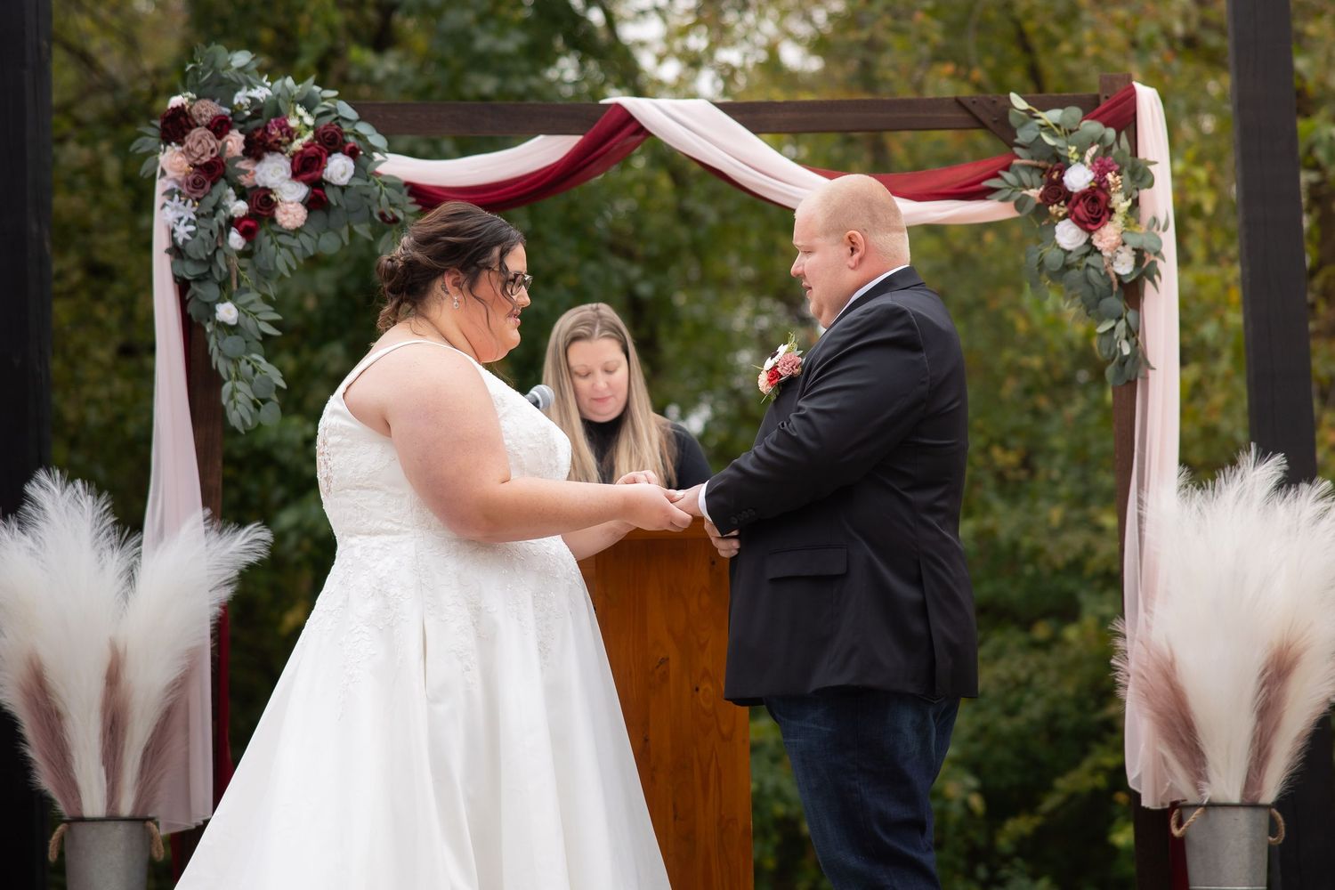 An outdoor wedding ceremony under a burgundy and floral decorated arch with pampas grass accents.