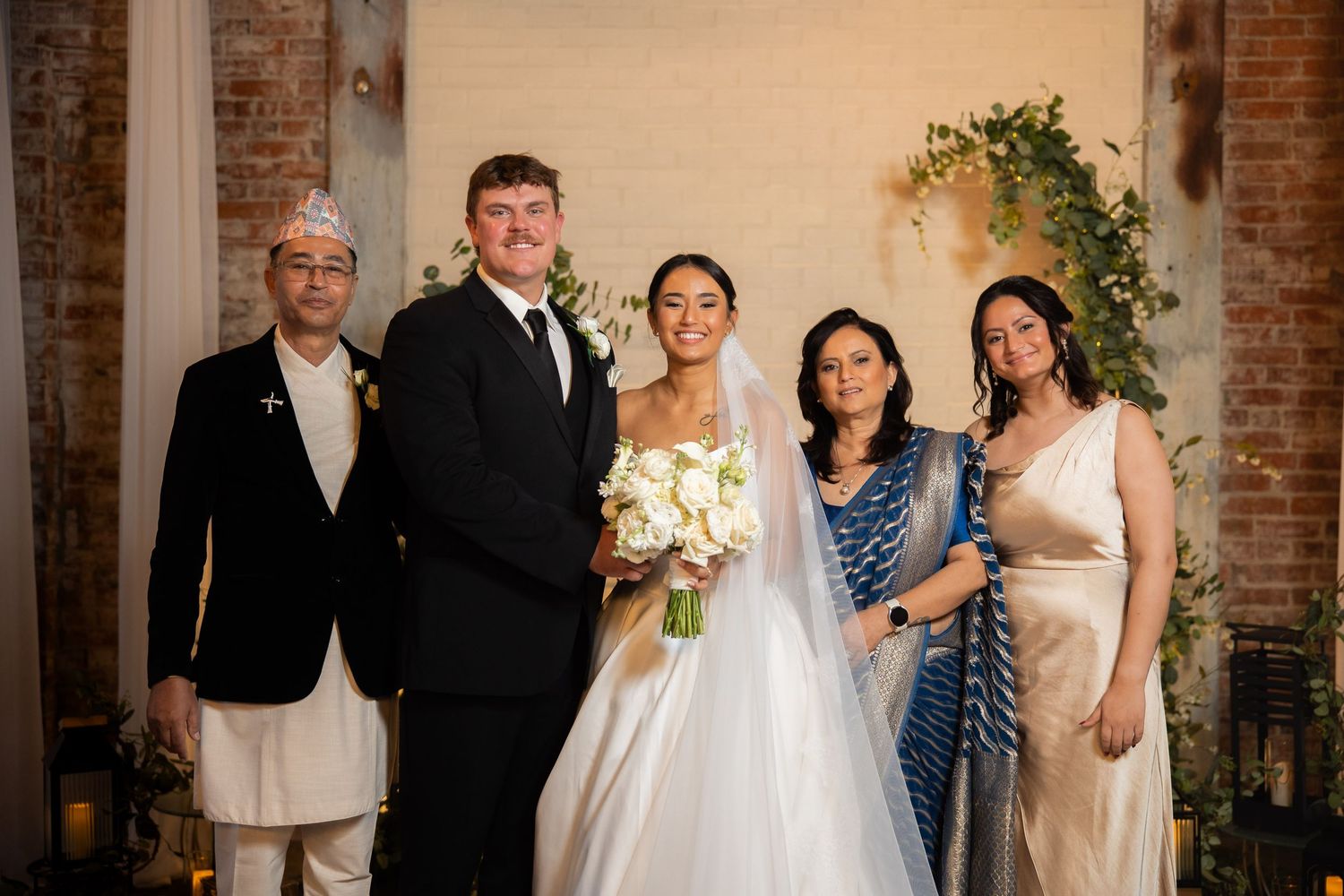 Wedding party lined up in formal attire against exposed brick wall under crystal chandeliers.