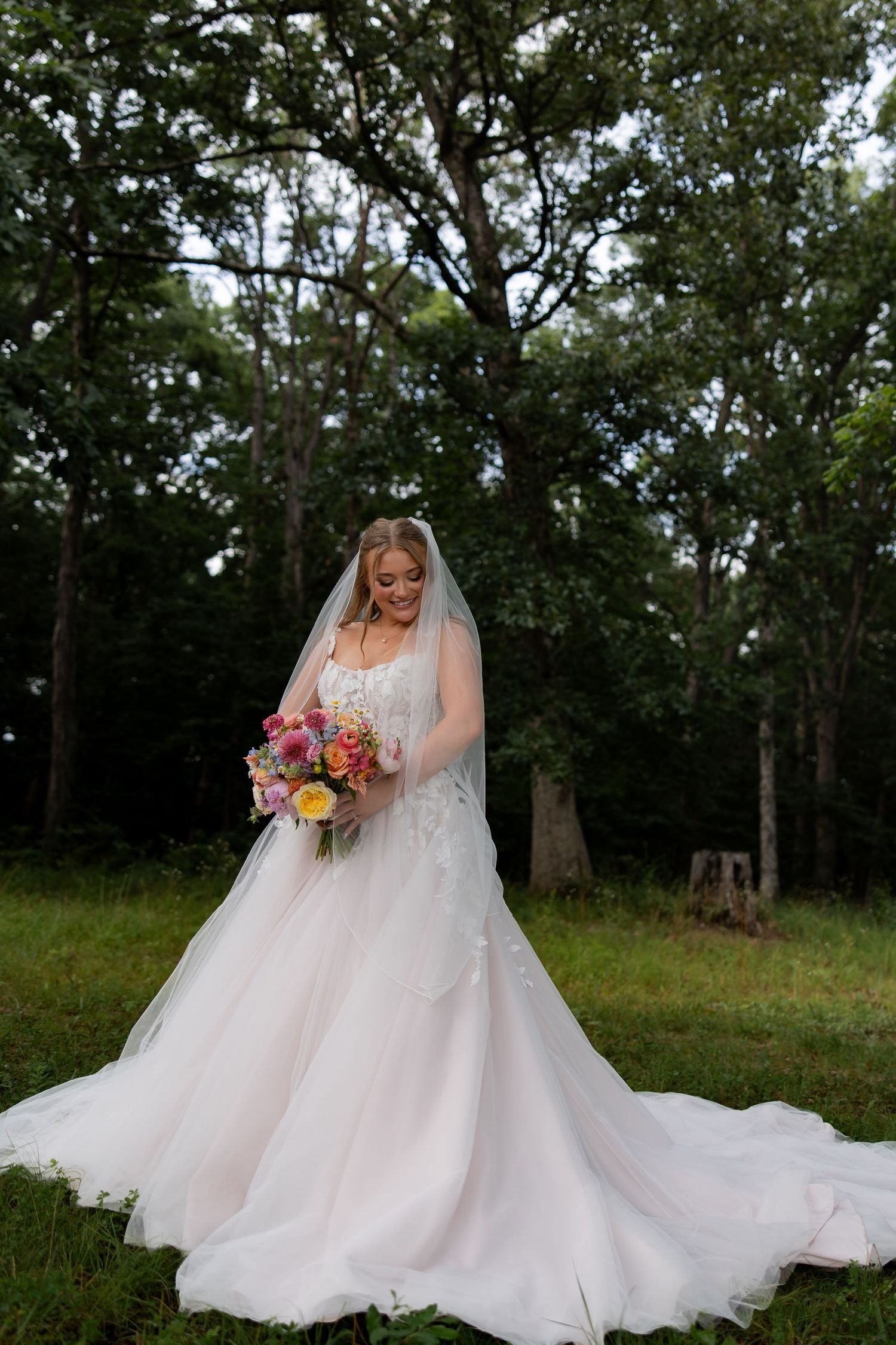 Bride in white lace gown with veil holds colorful bouquet outdoors among trees.