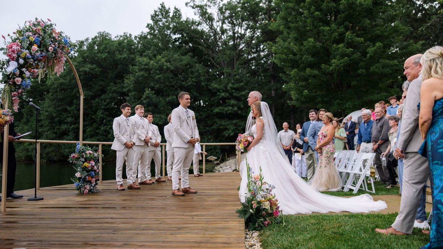 Bride walks toward groom on wooden dock ceremony with colorful floral arch and waterfront backdrop.