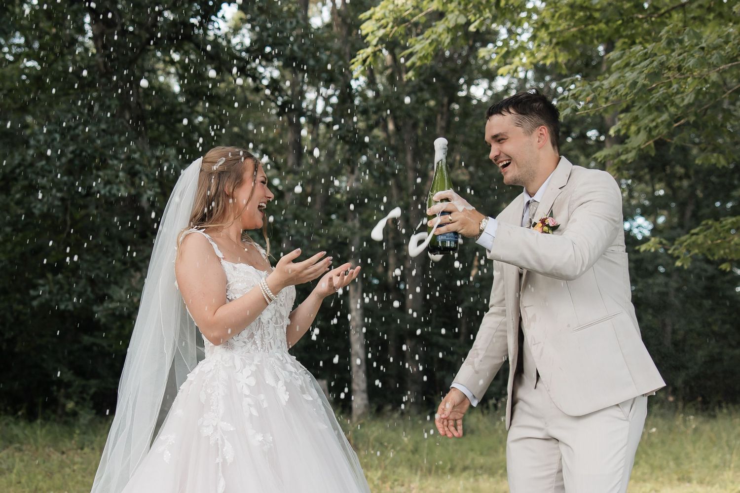 Bride and groom celebrating outdoors, popping champagne with confetti flying, surrounded by lush green trees.