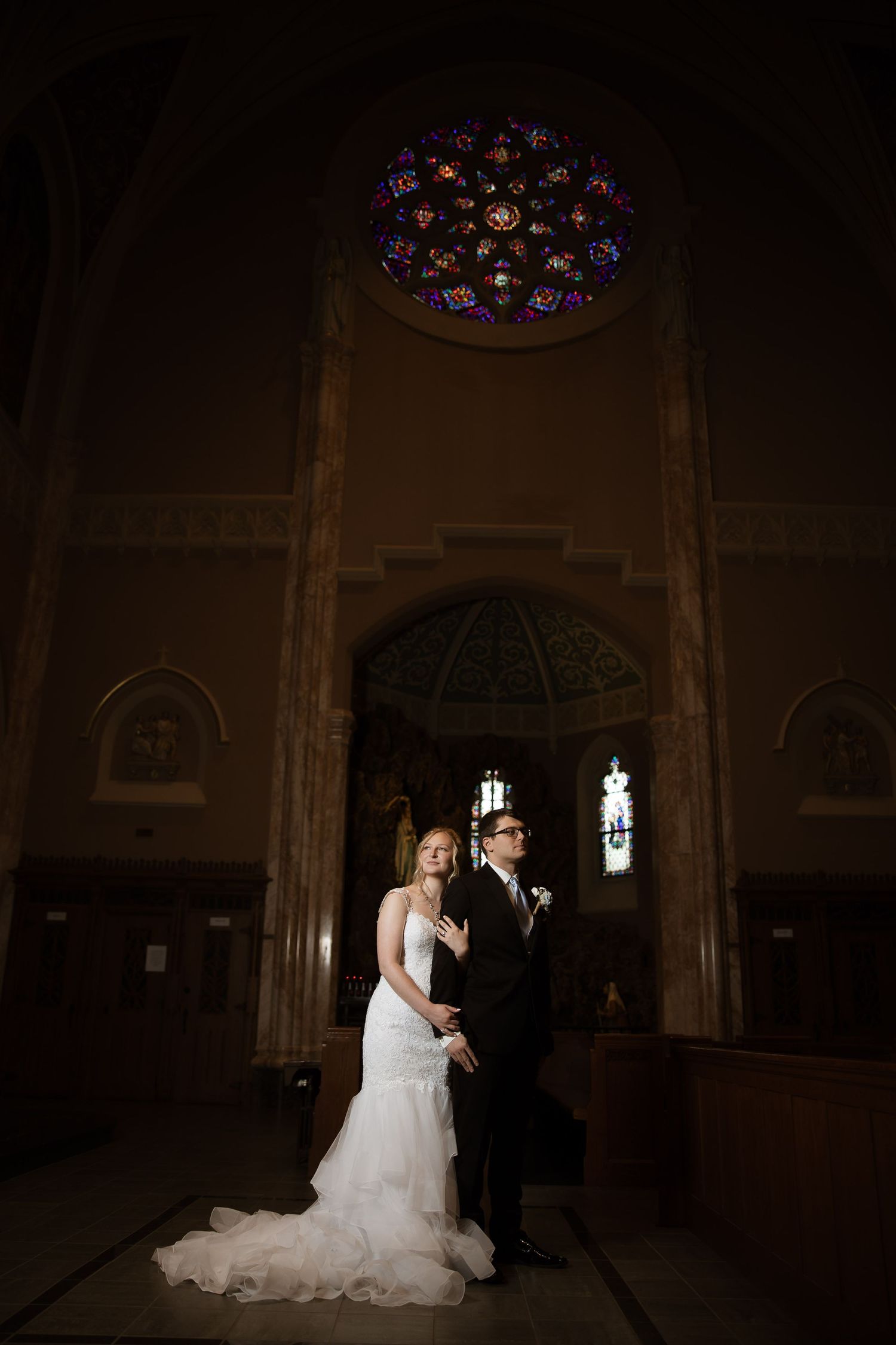 Dramatic silhouettes of a wedding couple inside a grand cathedral with stained glass windows and ornate architecture.