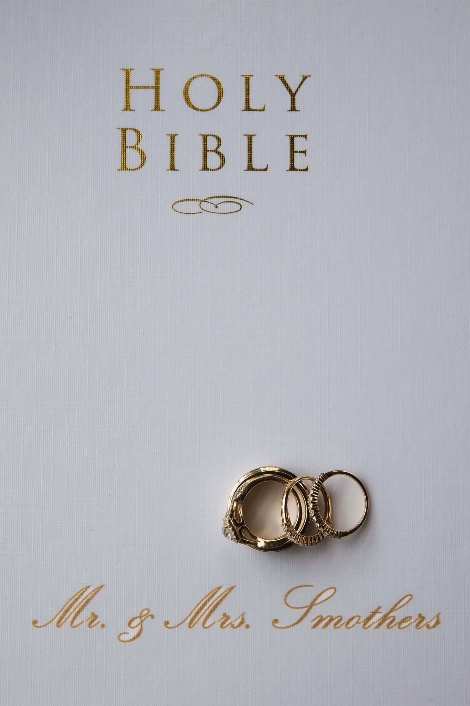Gold wedding rings photographed on a white Holy Bible cover with elegant script.