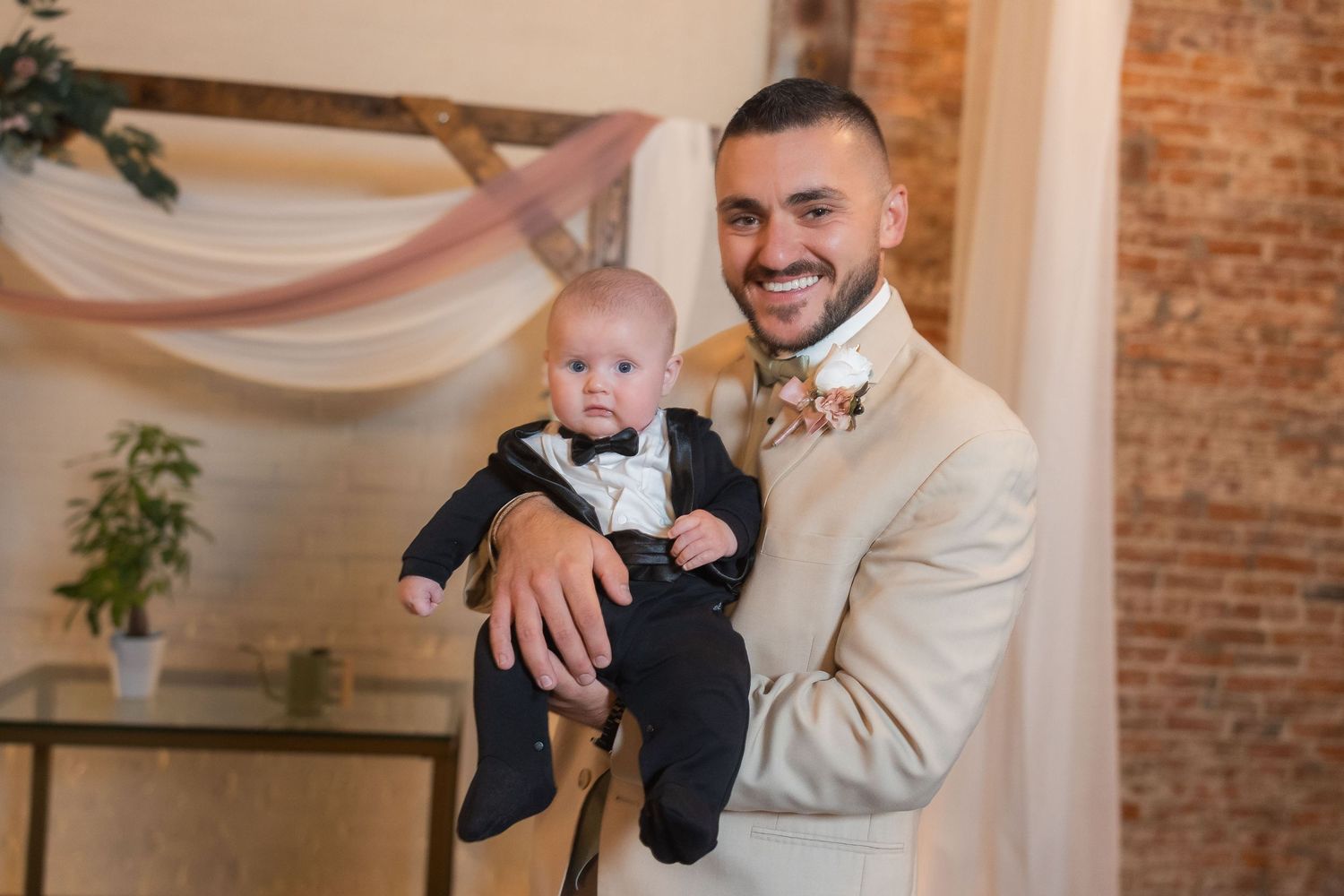 A formal event photo showing a baby in a tuxedo being held in an elegant indoor setting with exposed brick walls.
