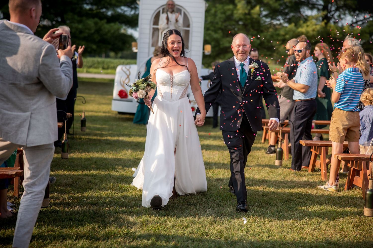 A joyful wedding ceremony outdoors on a sunny day with guests seated on wooden chairs along a grassy aisle.