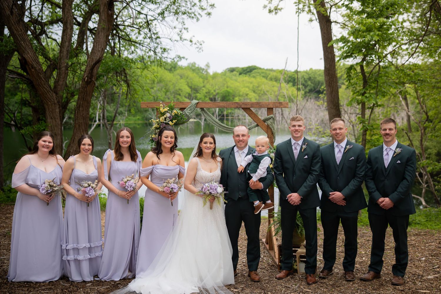Wedding party group photo by lakeside with bridesmaids in lavender gowns and groomsmen in dark green suits.