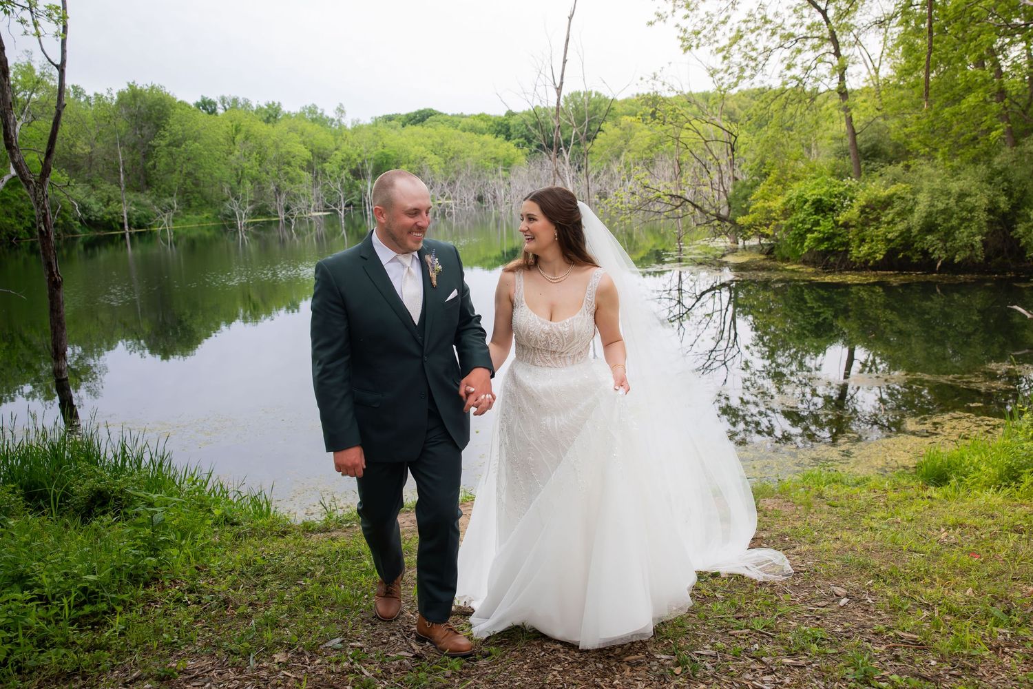 Smiling bride and groom walk hand in hand along a scenic lakeside on their wedding day.