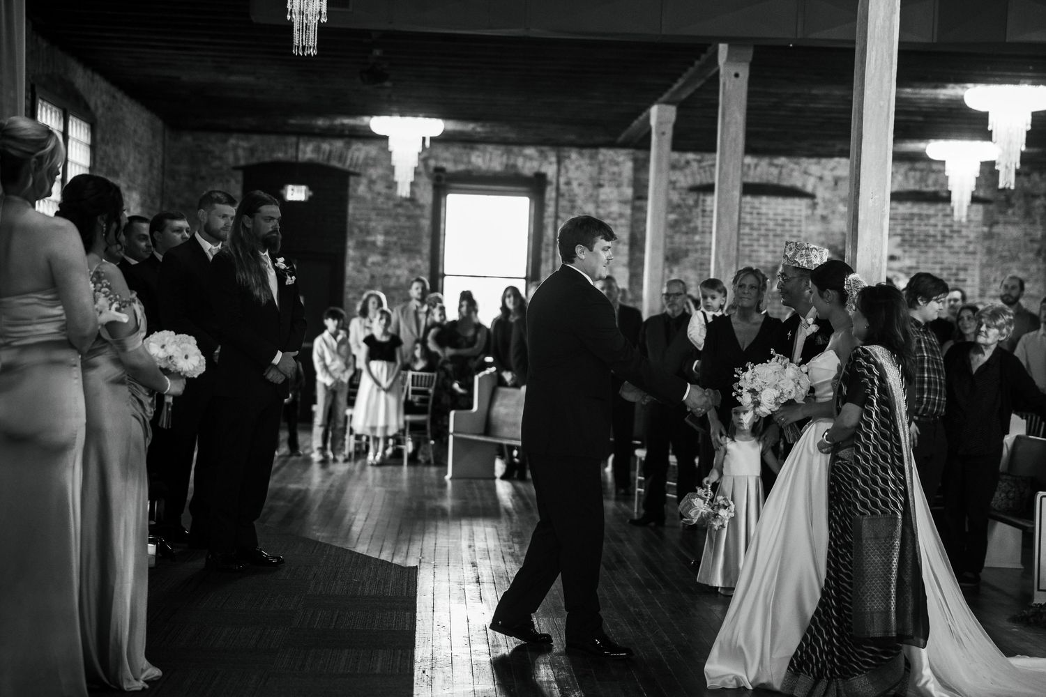 Black and white wedding ceremony photo showing groom walking bride down the aisle in elegant brick venue.