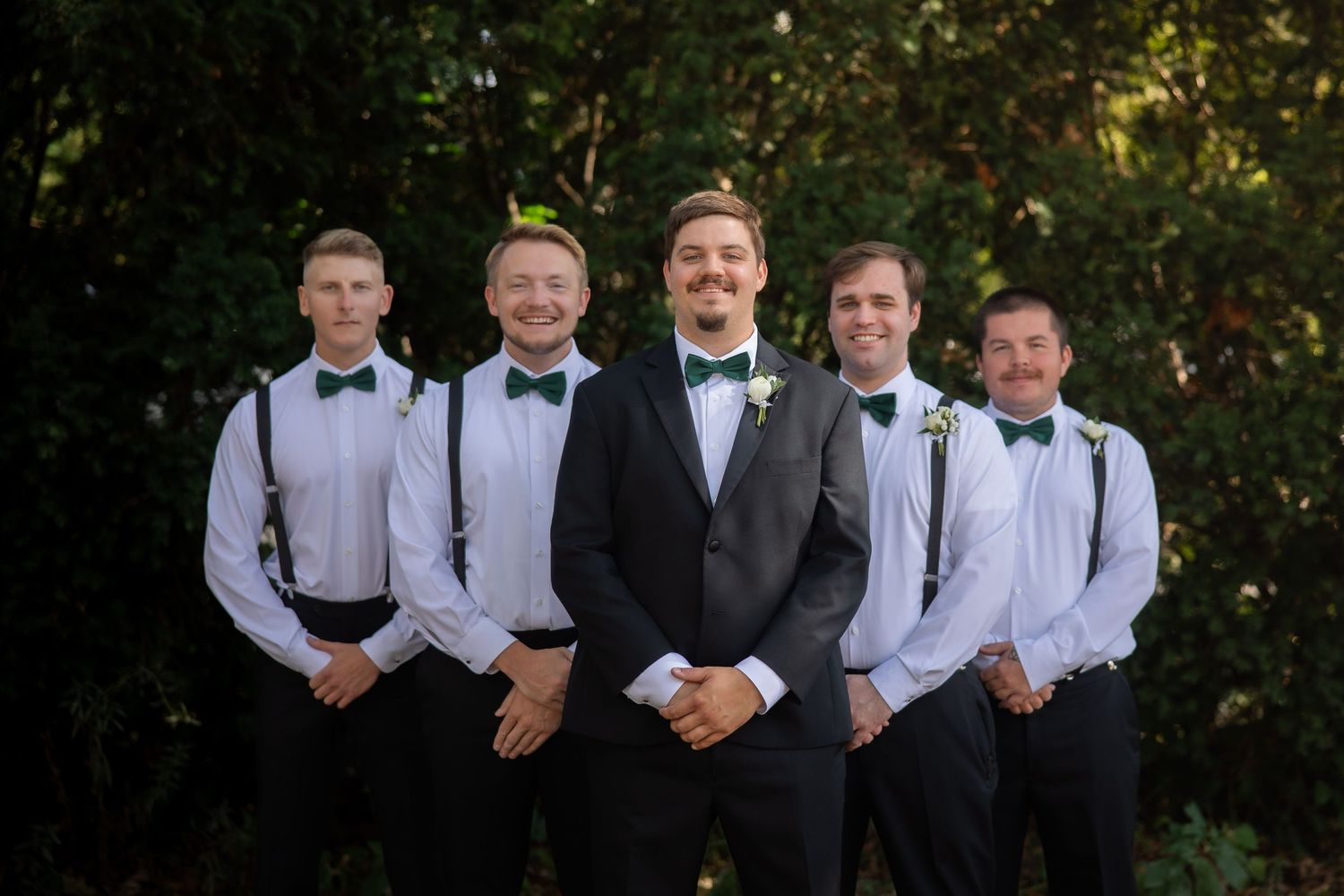 Groom in black tuxedo poses with four groomsmen wearing white shirts, green bow ties, and suspenders outdoors.