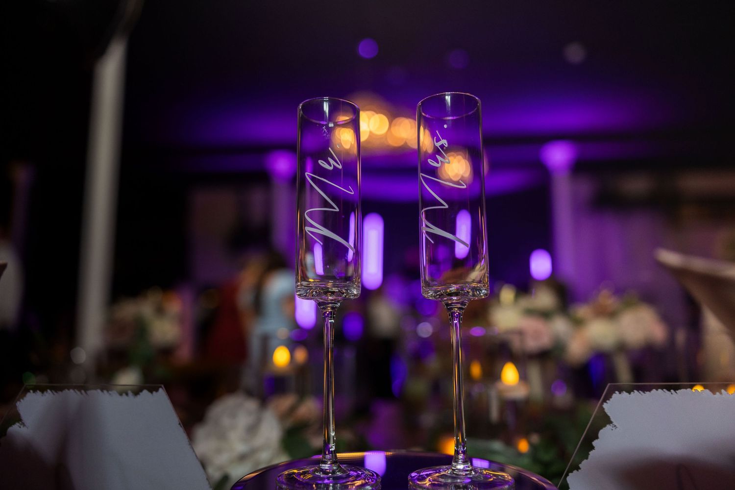 Wedding reception table settings with wine glasses illuminated by purple ambient lighting.