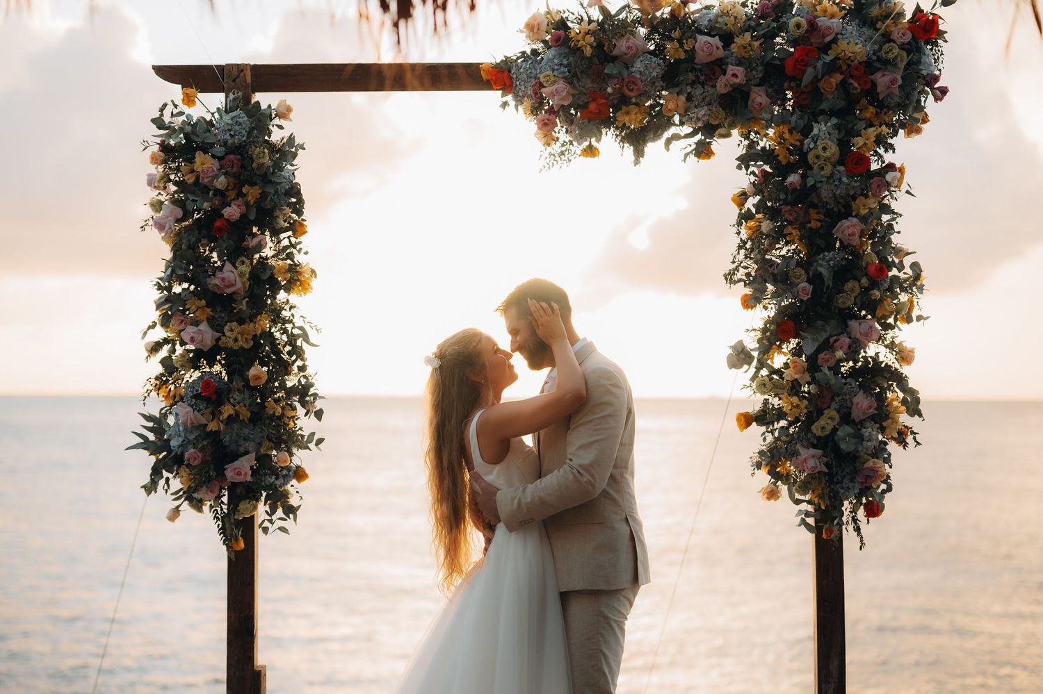 Wedding ceremony setup with floral arch and couple silhouetted against sunset sky.