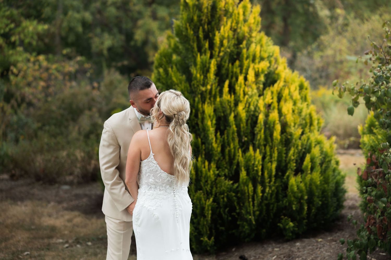 Romantic first look wedding photos captured in garden setting with evergreen shrubs.