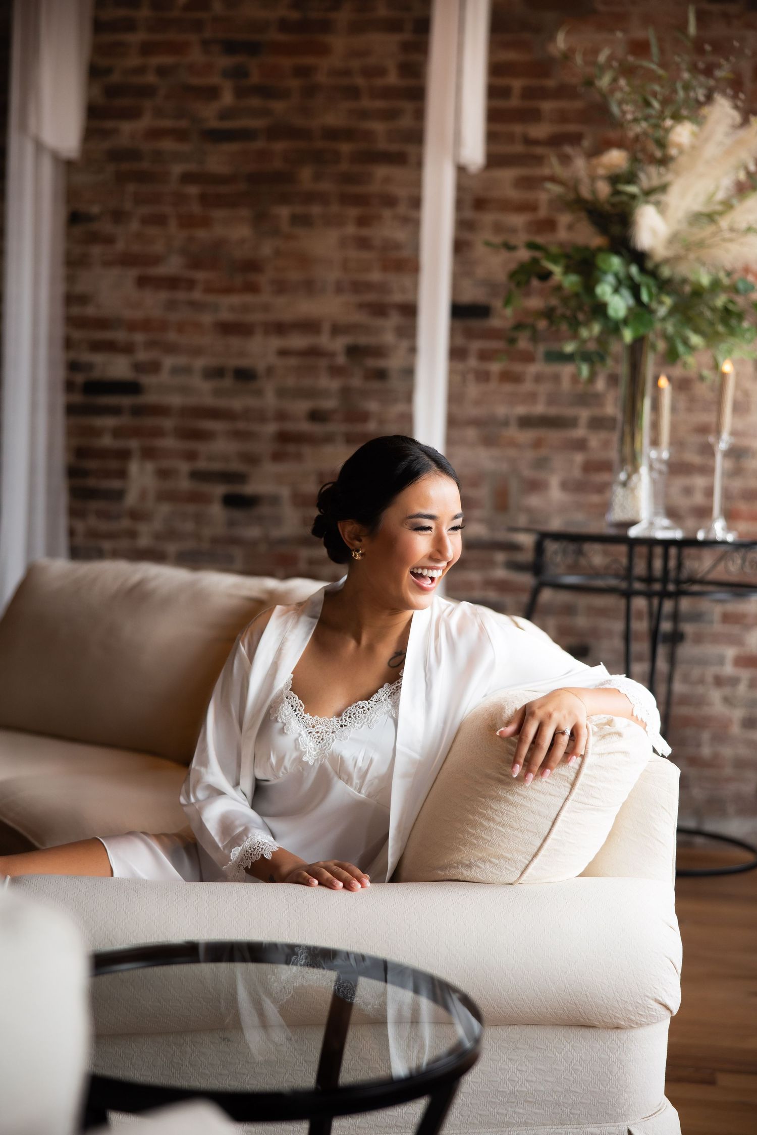 A bride in a white robe relaxes on a beige couch against exposed brick walls with elegant floral decor.