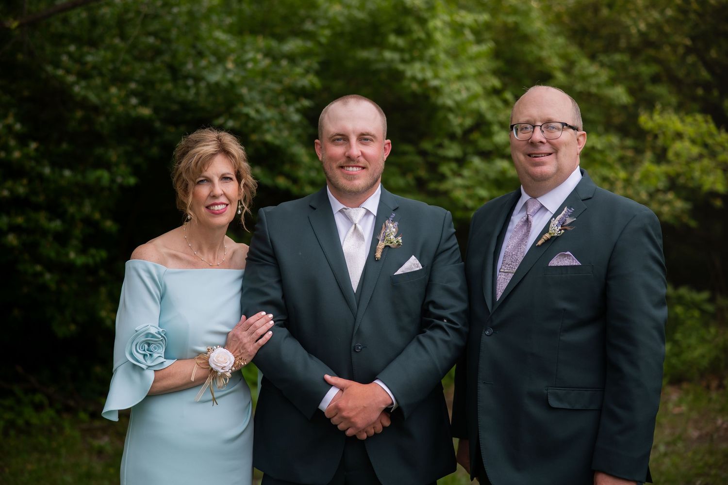 Groom in gray suit poses with smiling parents in formal attire outdoors at a wedding celebration.