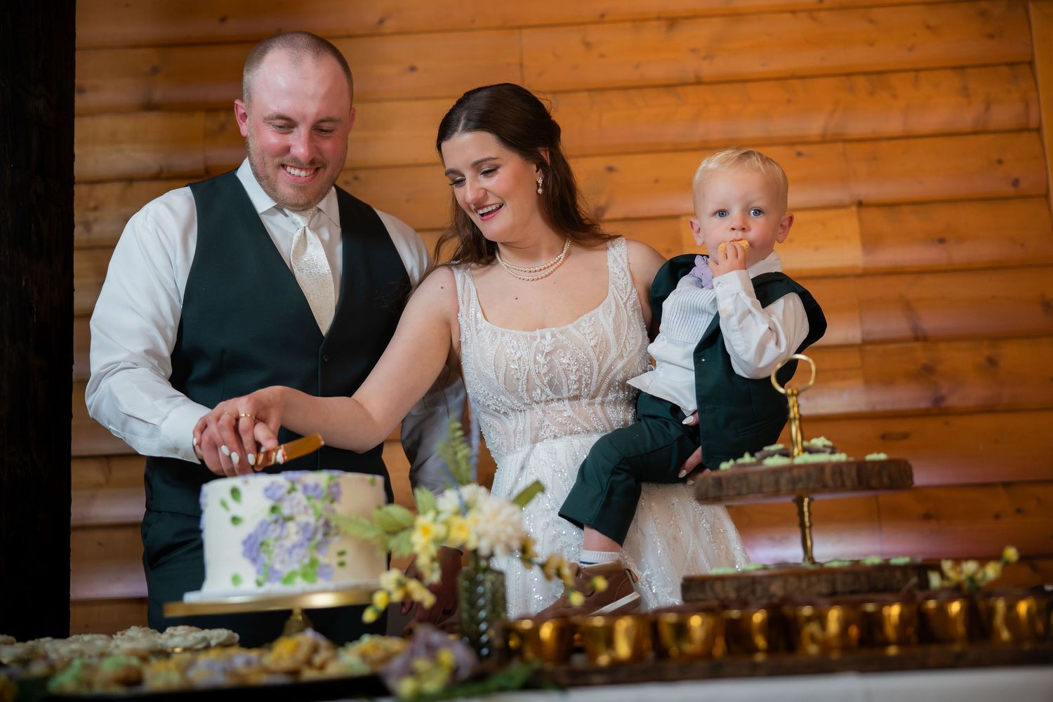 Smiling bride and groom cut wedding cake while holding young baby in tuxedo at rustic reception.