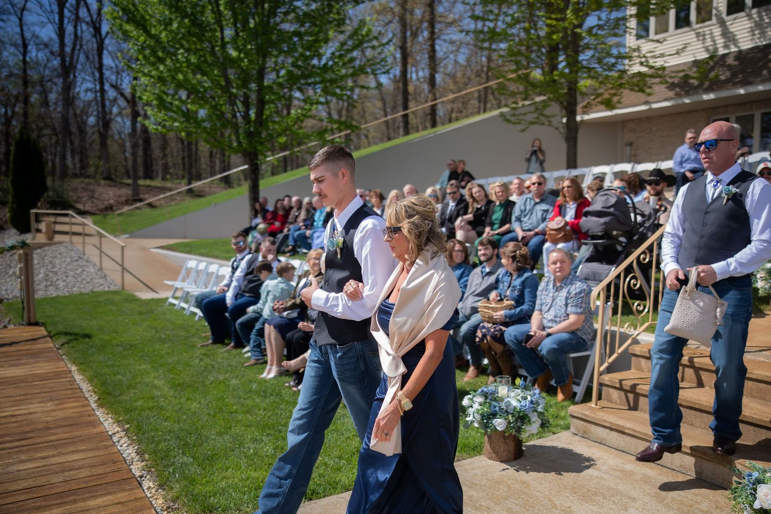 Wedding guests seated outdoors watch couple walk down aisle at scenic garden ceremony venue.