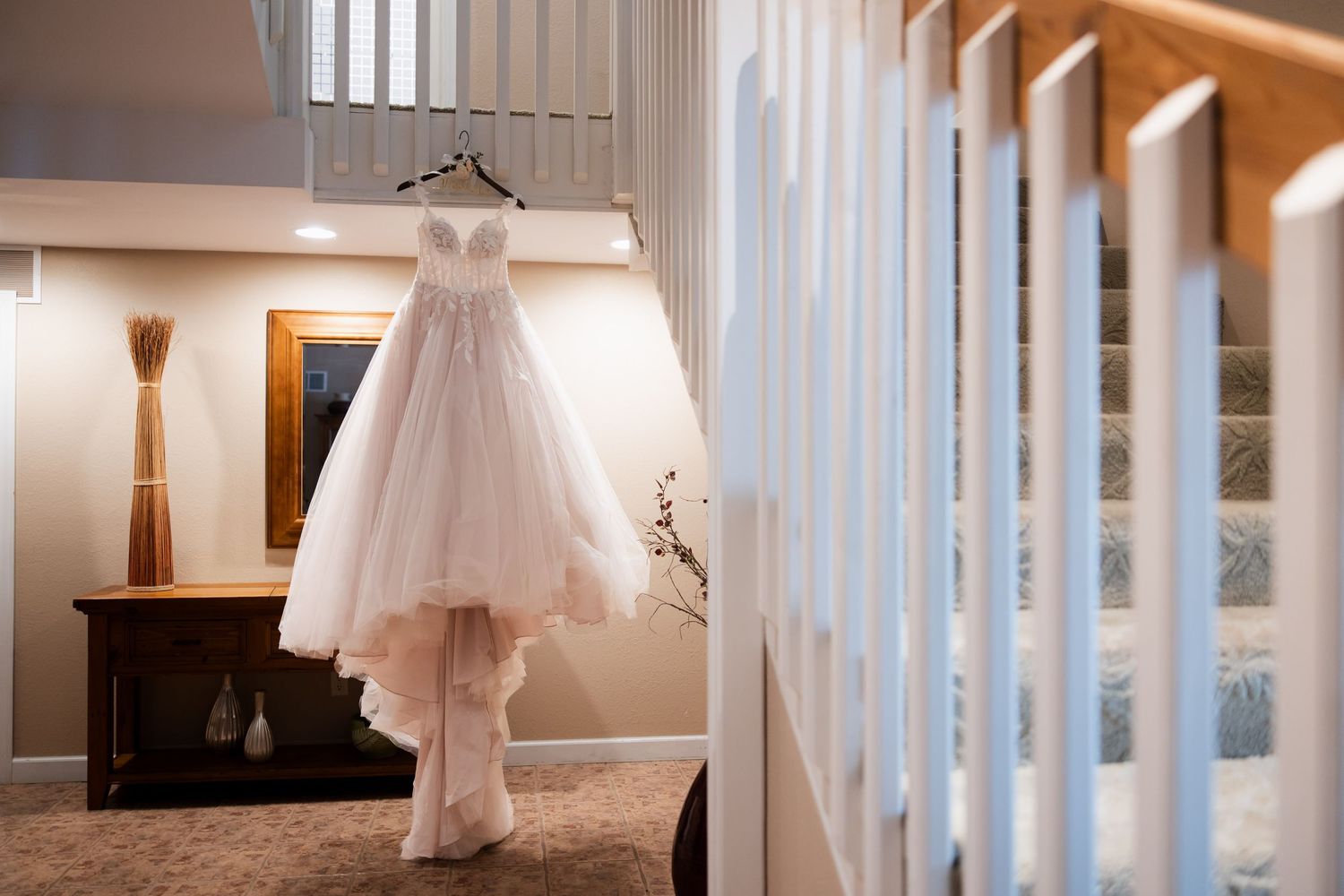 Elegant wedding dress hanging on staircase railing in warmly lit home interior.