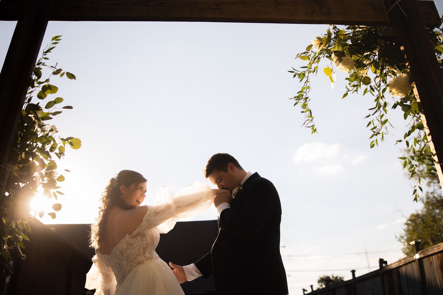 Bride and groom exchanging vows under a sunlit floral arch with golden backlight.