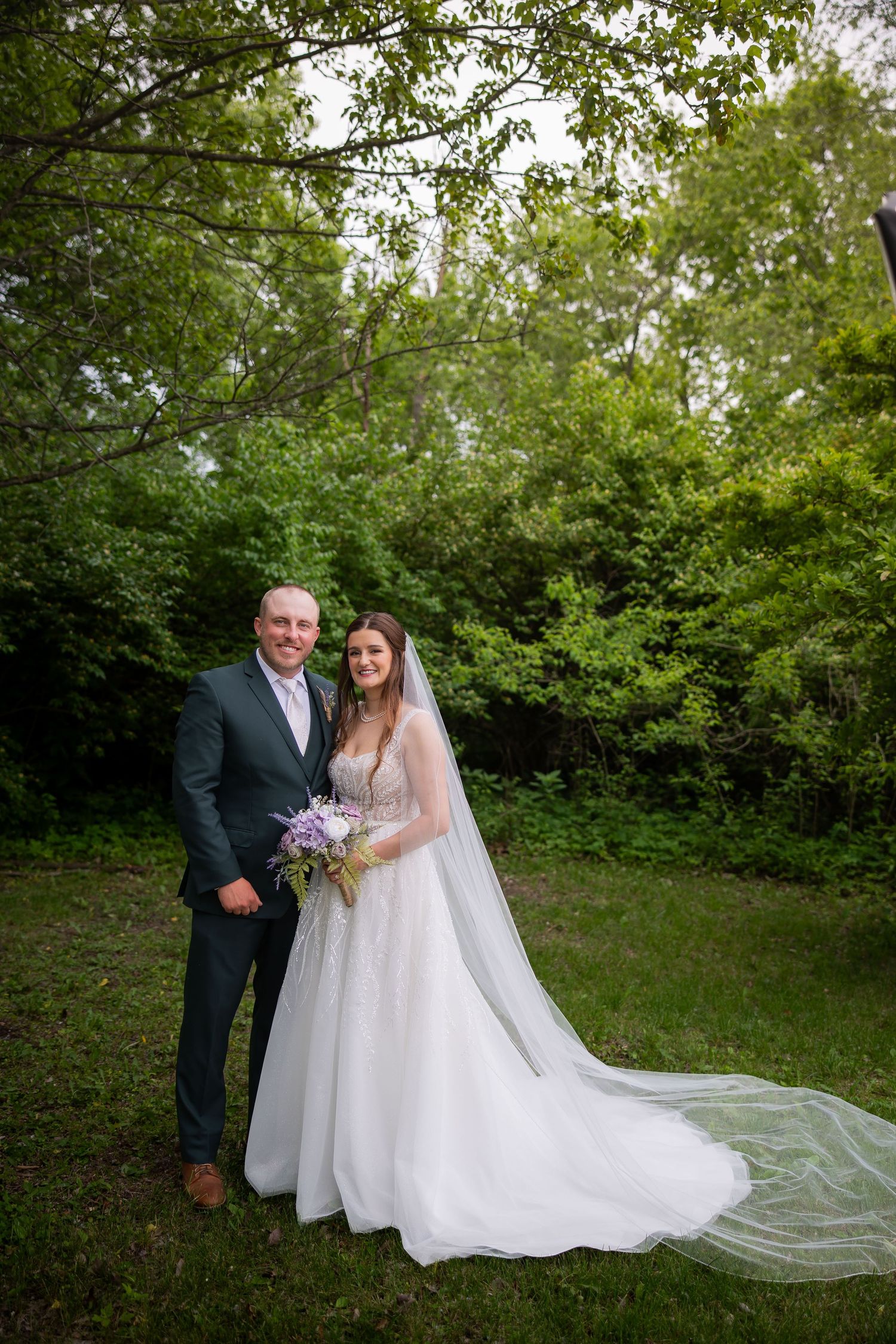 Bride and groom pose together in lush green garden, bride wearing elegant white gown with long veil holding purple bouquet.