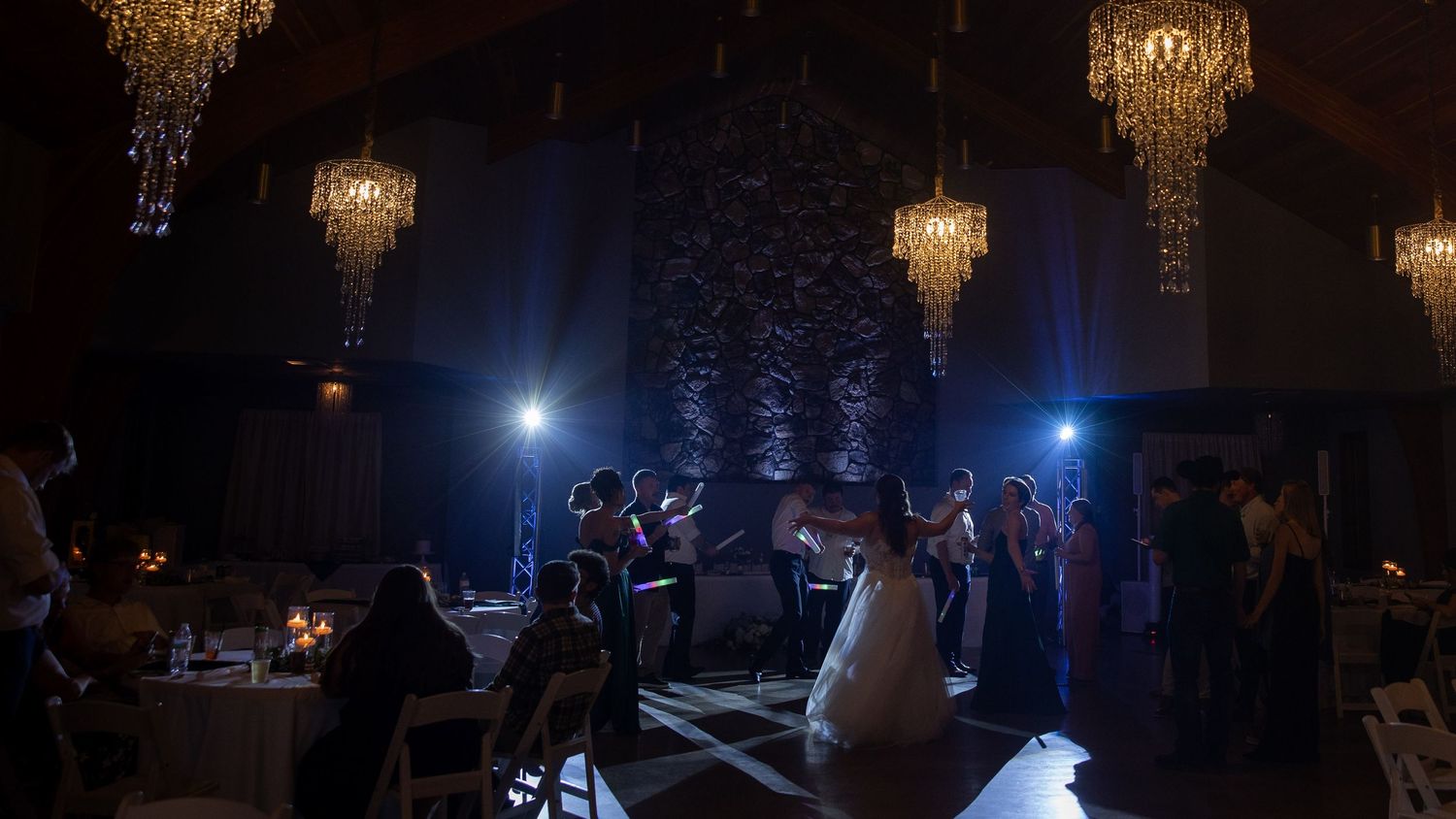 Elegant wedding reception with chandeliers, bride on dance floor, guests seated, blue stage lighting in dark venue.