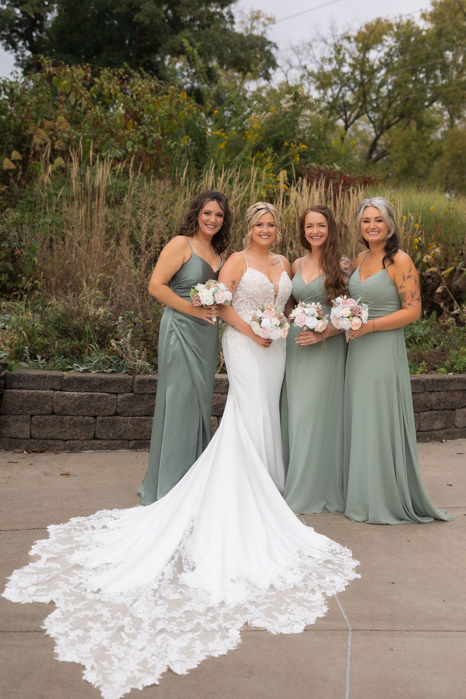 Bride in lace gown with long train poses with bridesmaids wearing sage green dresses in garden setting.