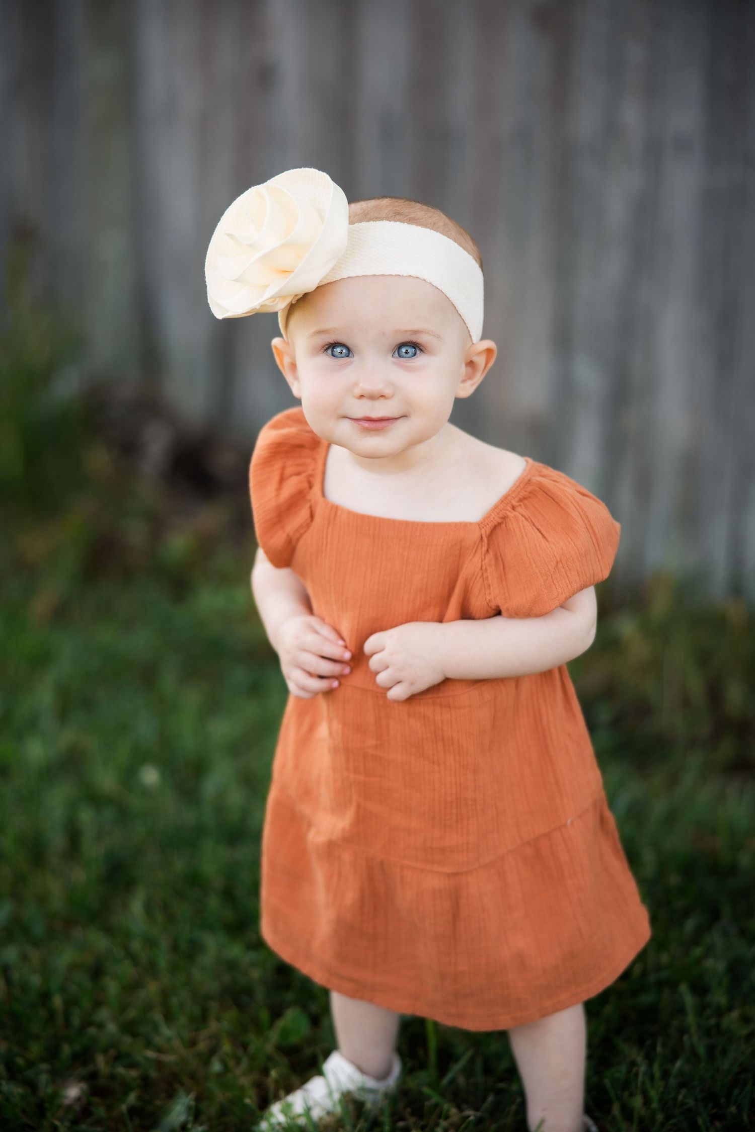 Baby in orange dress sits on grass in front of wooden fence during outdoor photo session.