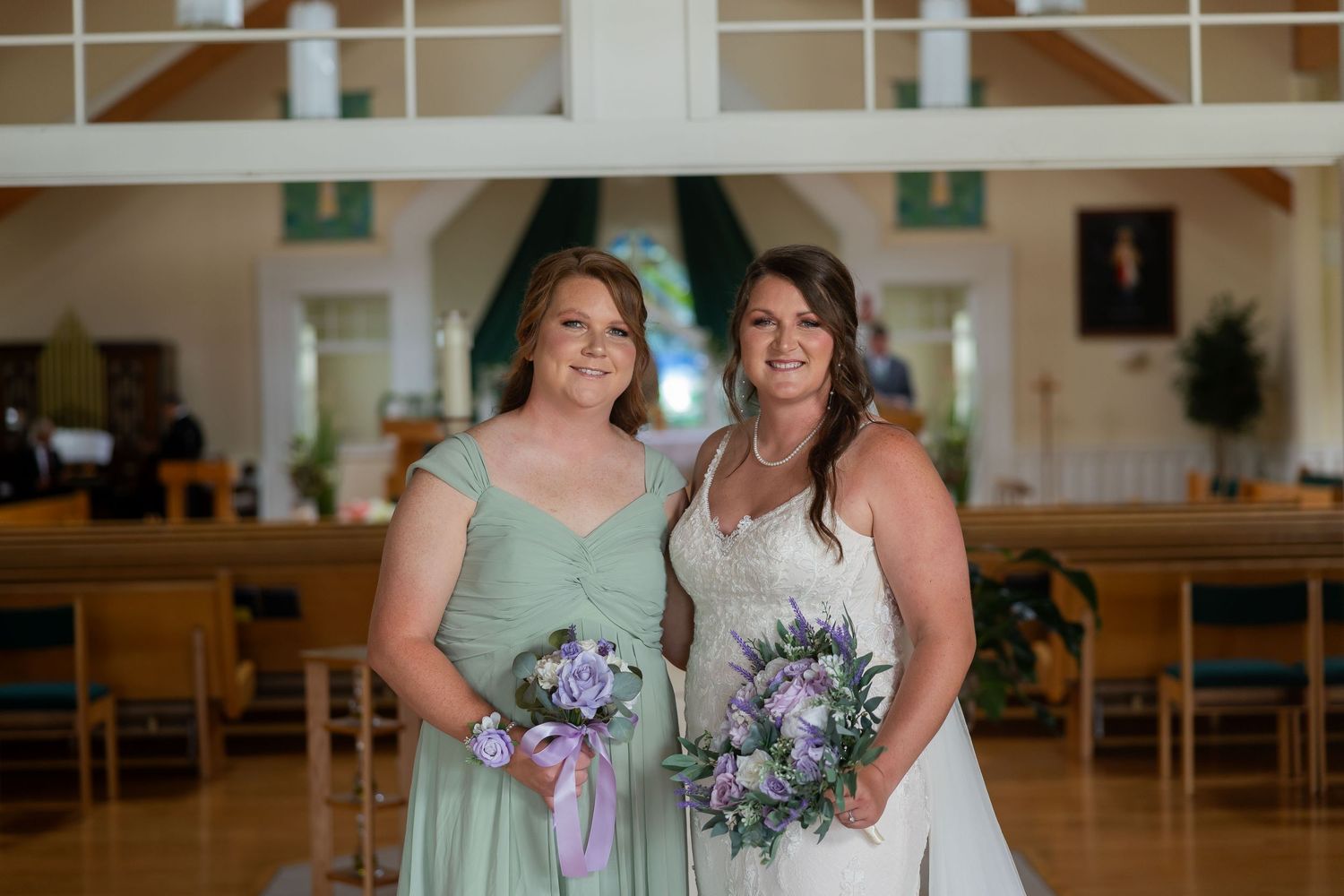 Bride in white gown and bridesmaid in mint green dress holding purple bouquets inside a church.