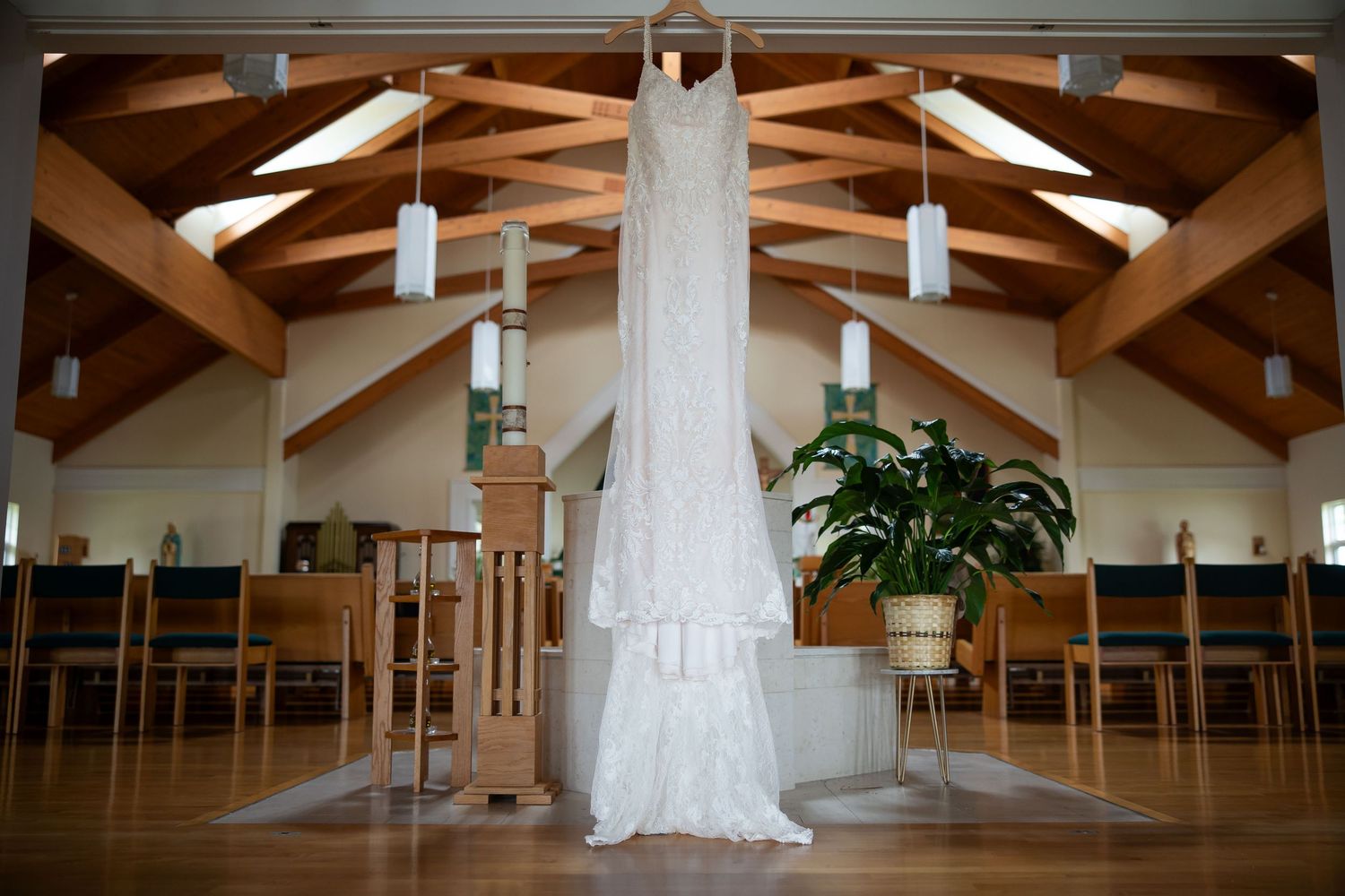 Elegant white wedding dress hanging in a church with wooden beam ceilings and pendant lights.