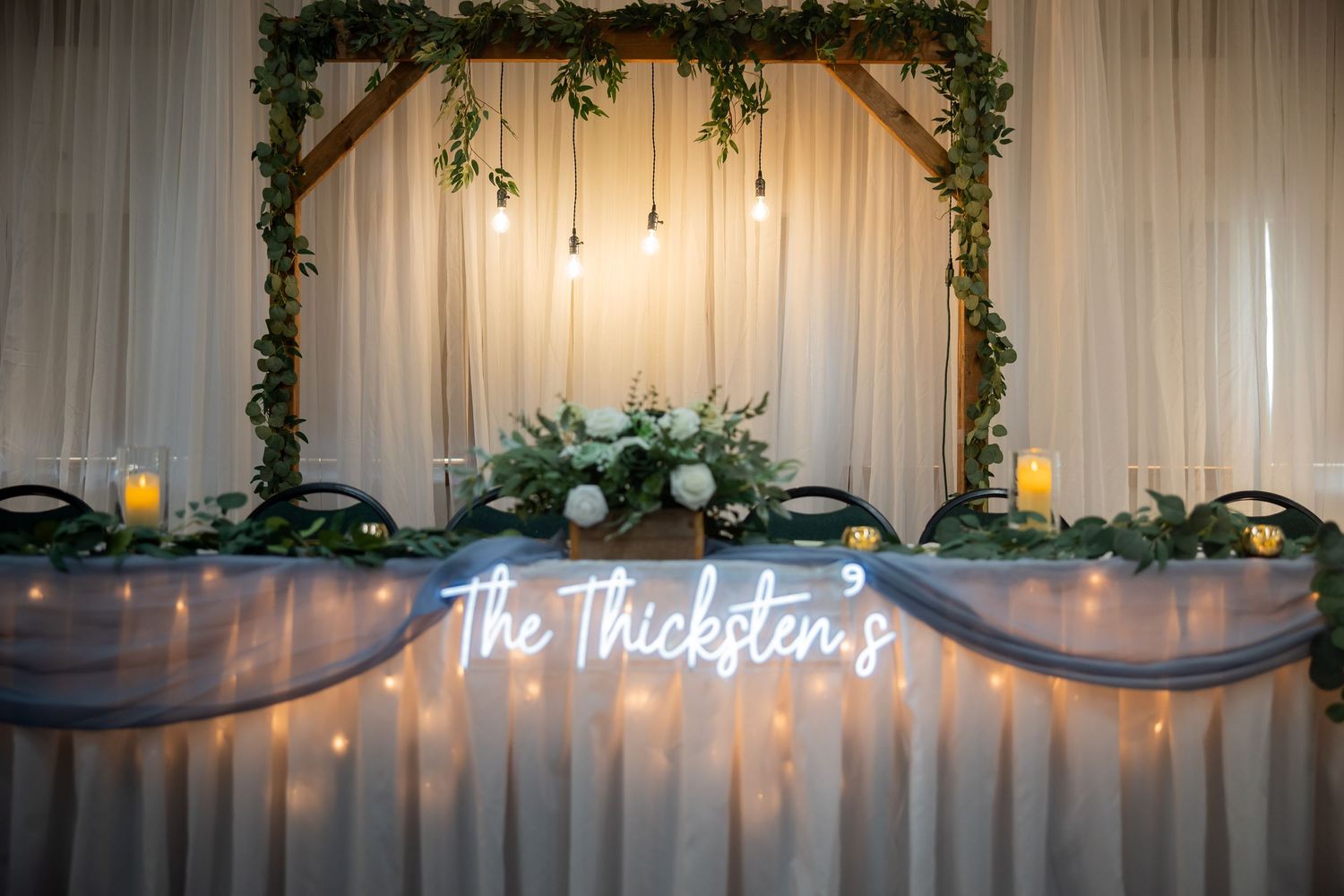 Wedding reception head table decorated with greenery arch, fairy lights, and The Thicksten's neon sign.