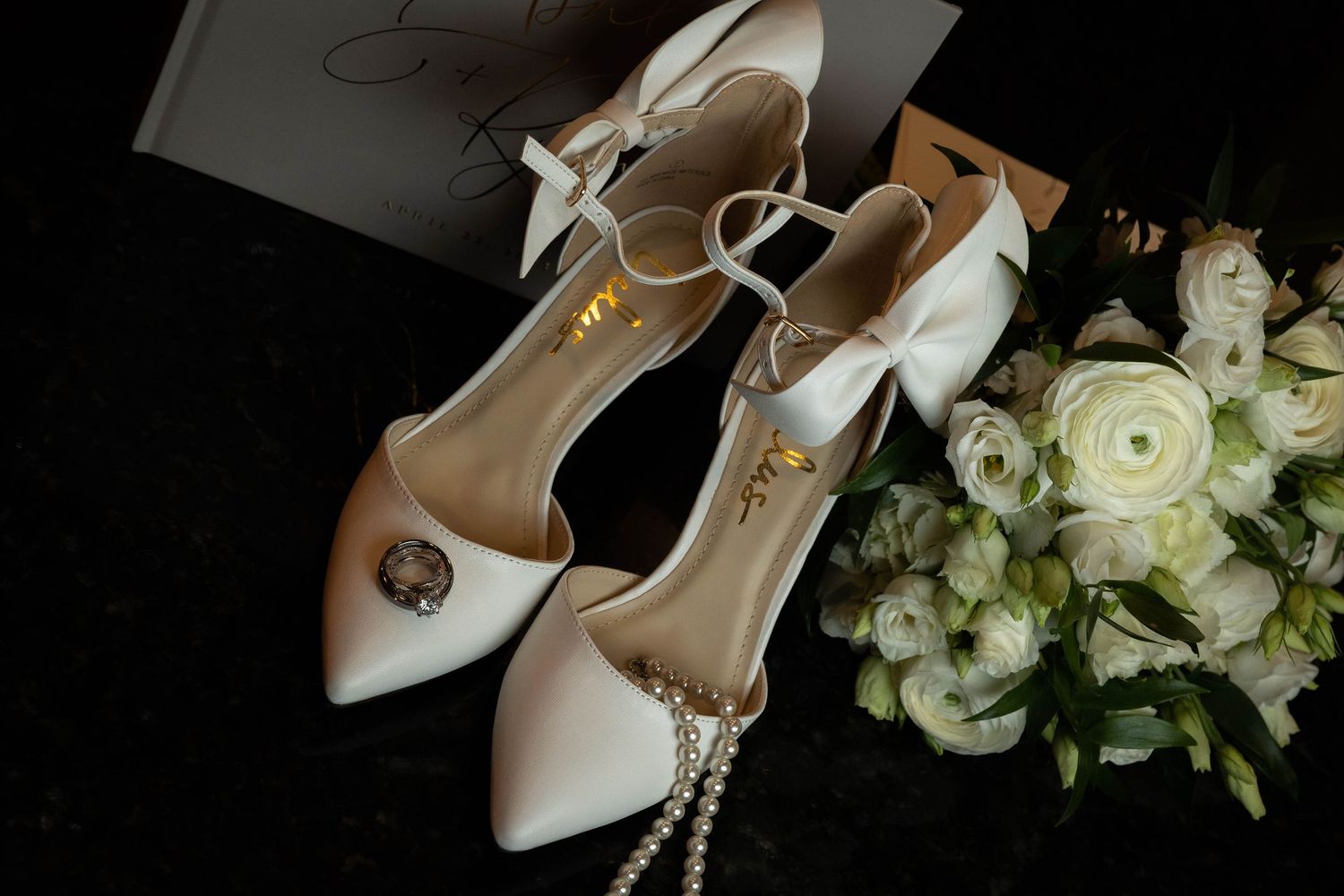 Elegant white satin bridal heels with jewel detail and ankle strap beside white roses and pearl necklace.