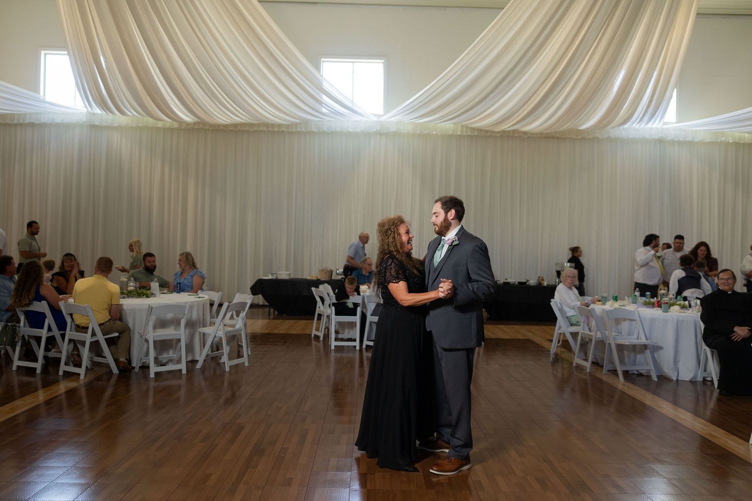 Couple sharing a romantic dance at a wedding reception in an elegant white draped venue with guests seated at round tables.