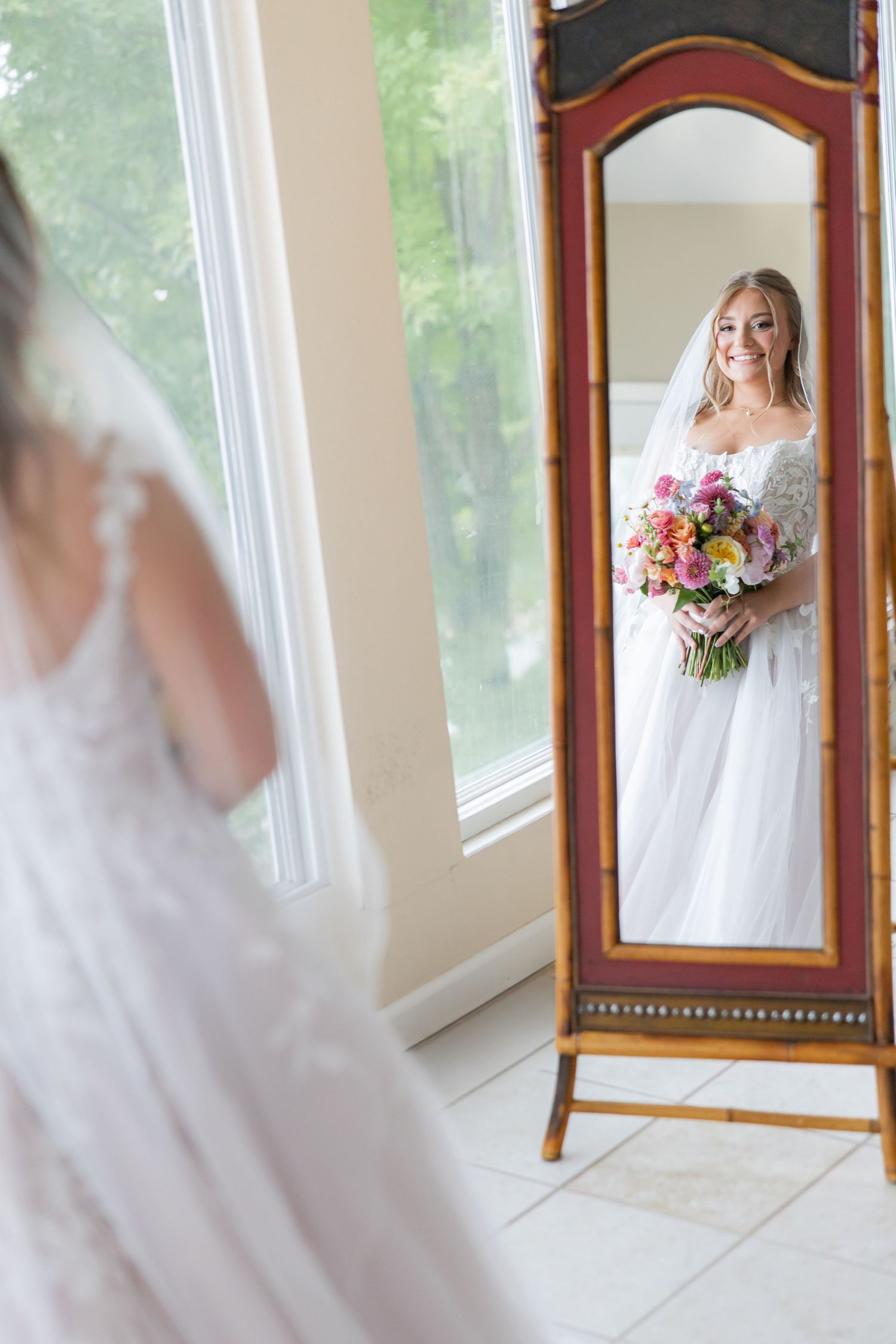Bride in white gown holding colorful bouquet admires her reflection in an ornate standing mirror.