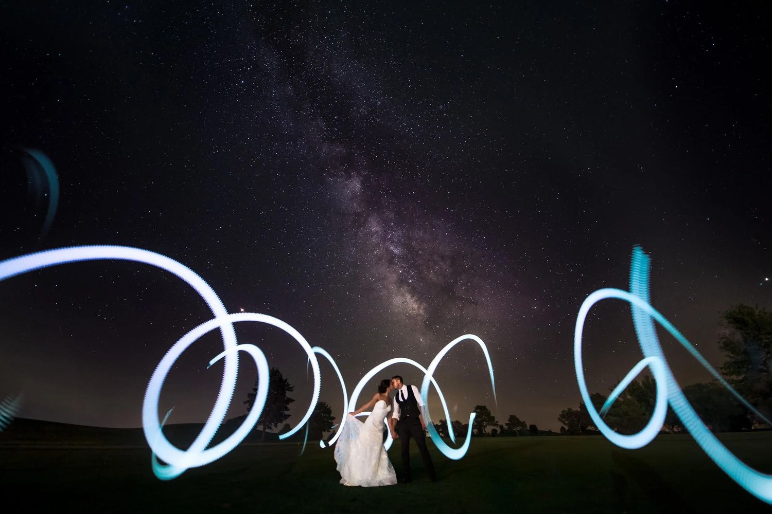 Wedding couple under Milky Way night sky with glowing light painting swirls creating magical long-exposure photography art.