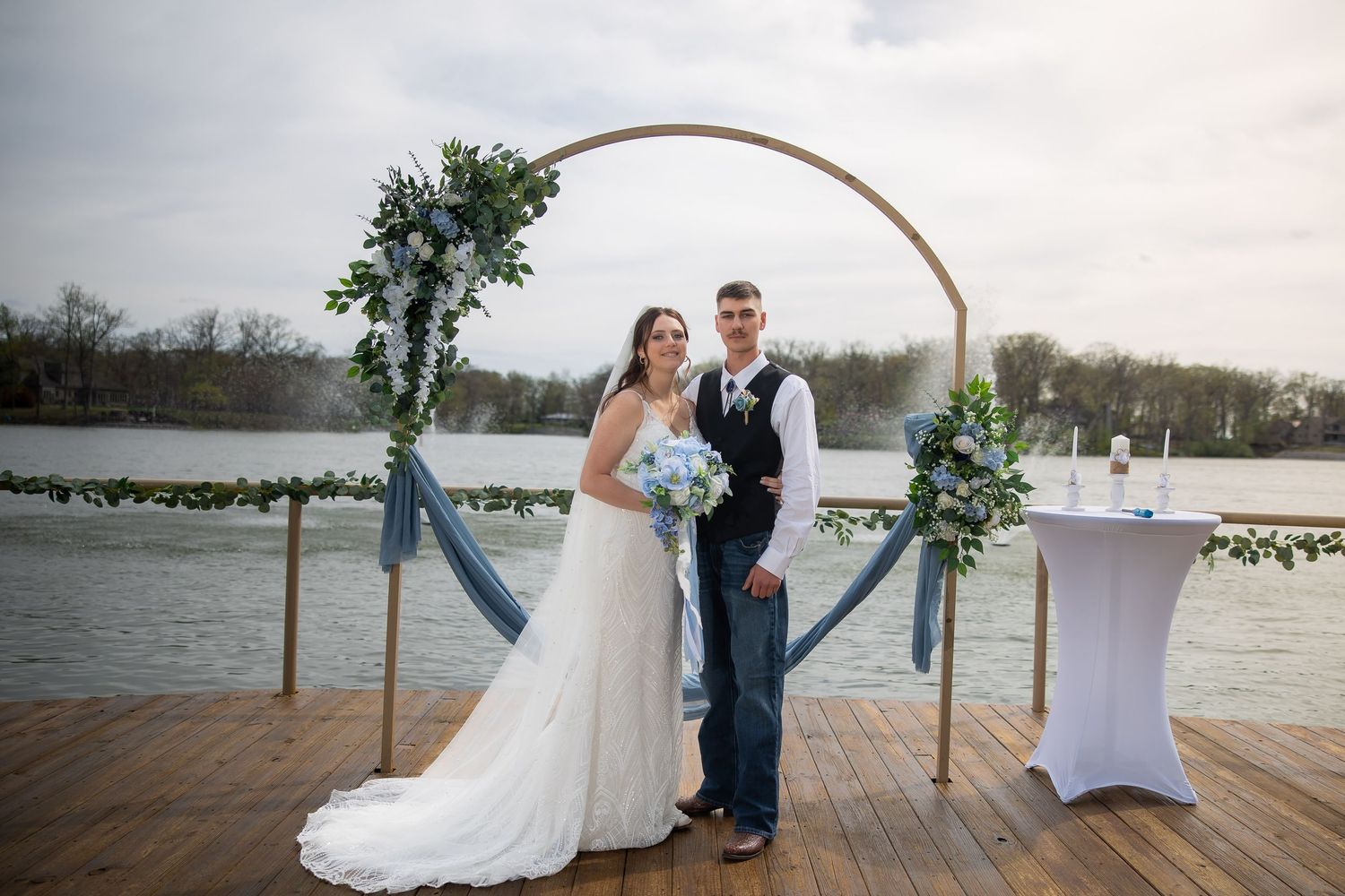 Bride and groom stand beneath a circular greenery arch on a lakeside dock.