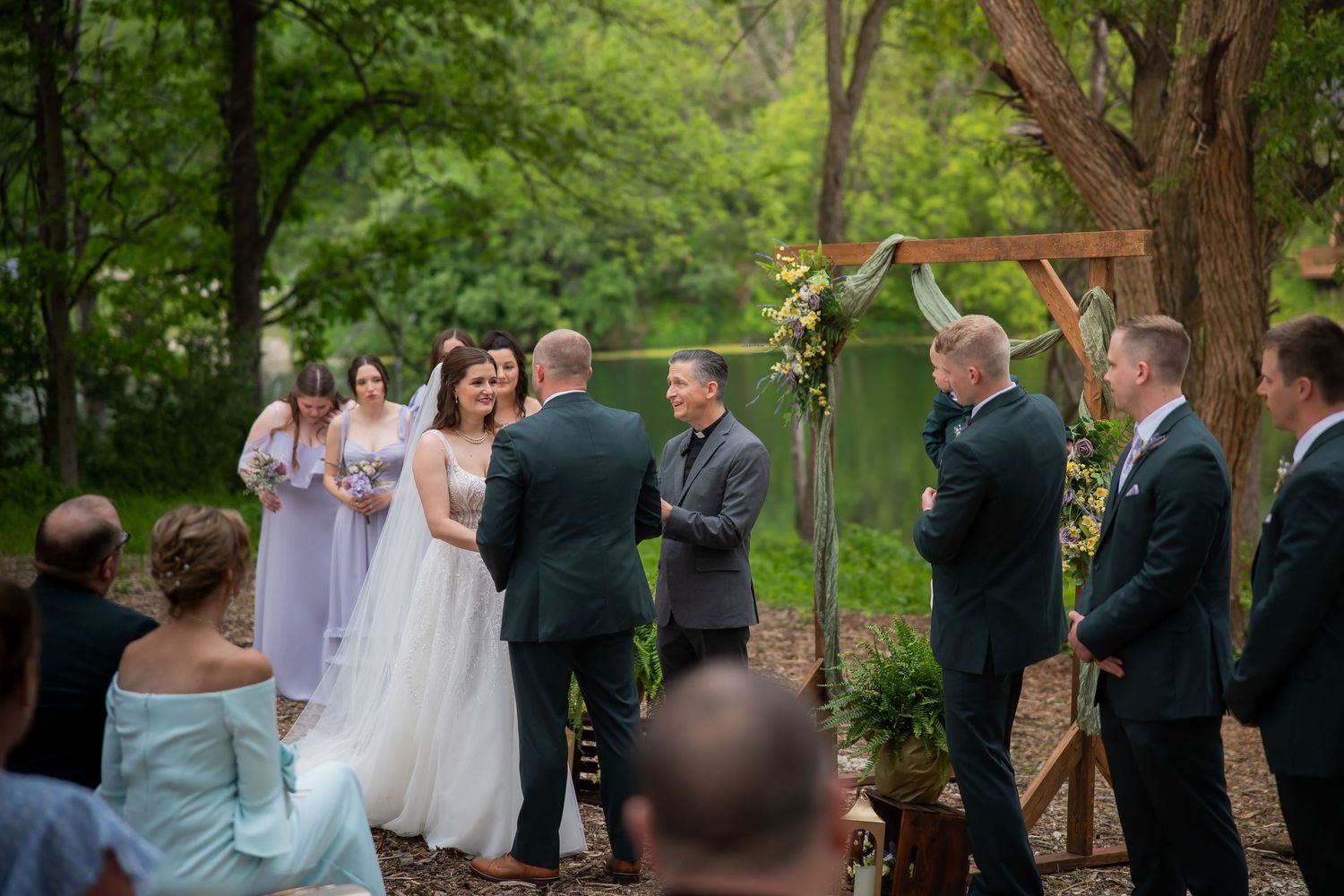 Bride and groom exchange vows at rustic outdoor woodland wedding ceremony with wedding party and guests.