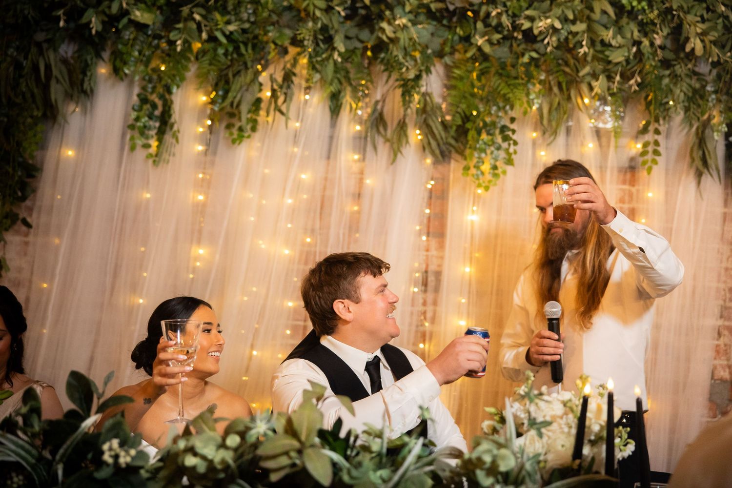Wedding celebration with guests toasting under hanging greenery and warm lighting.