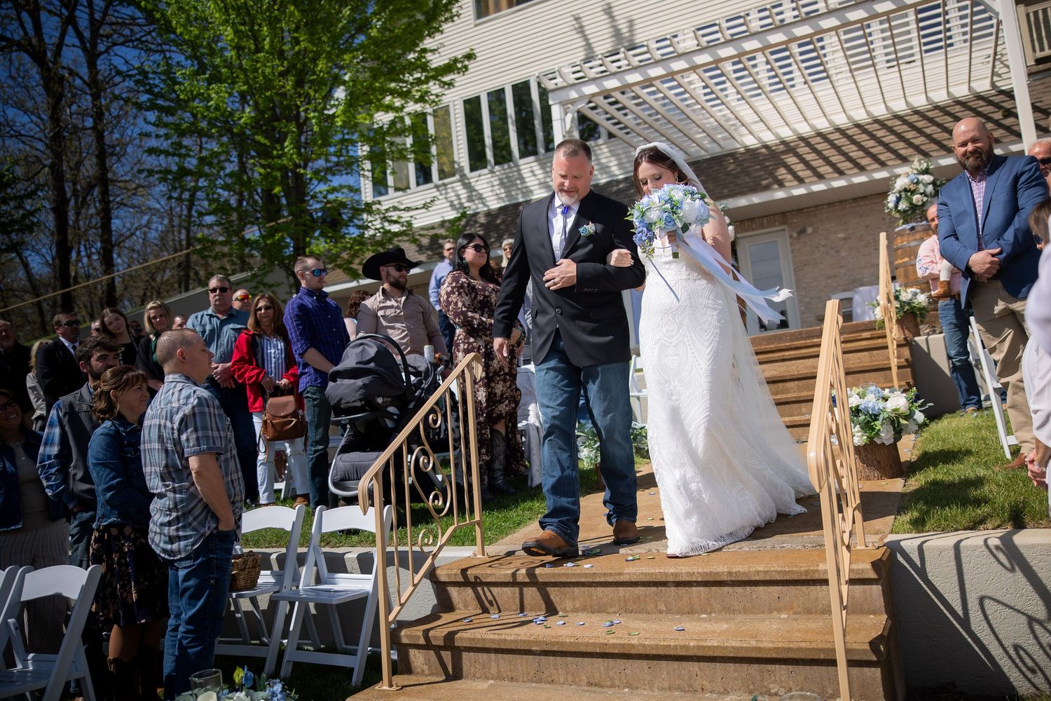 Bride walks down outdoor wedding aisle steps with escort, guests seated beside decorated wooden staircase.