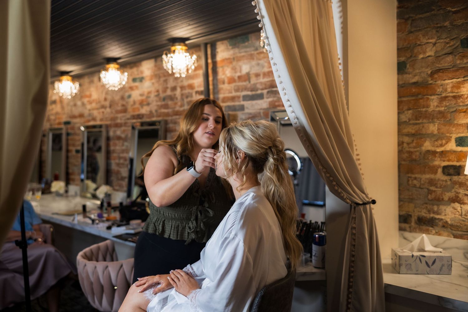 Professional makeup artist applies cosmetics to client in a rustic brick-walled salon setting.