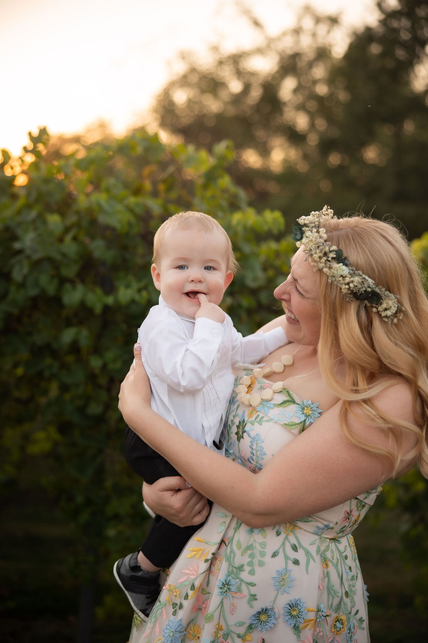 A sweet outdoor moment at sunset with a baby in white and a woman in a floral dress wearing a flower crown.