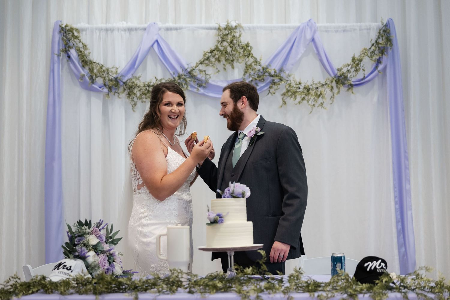 Smiling bride and groom cut their white wedding cake at elegant reception with purple floral backdrop.