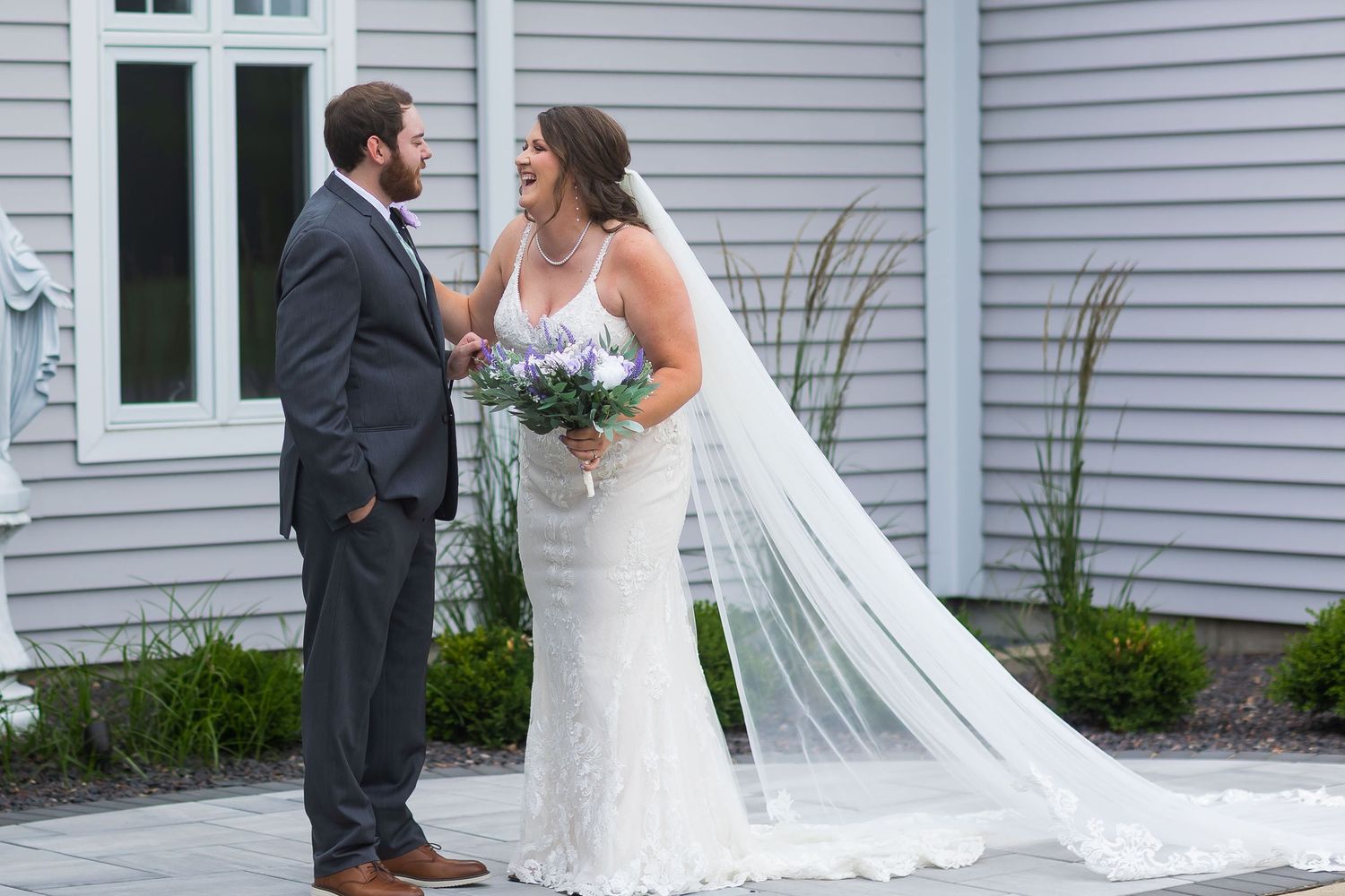 Bride and groom share a romantic moment outside a gray building, bride holding a lavender bouquet with a flowing veil.